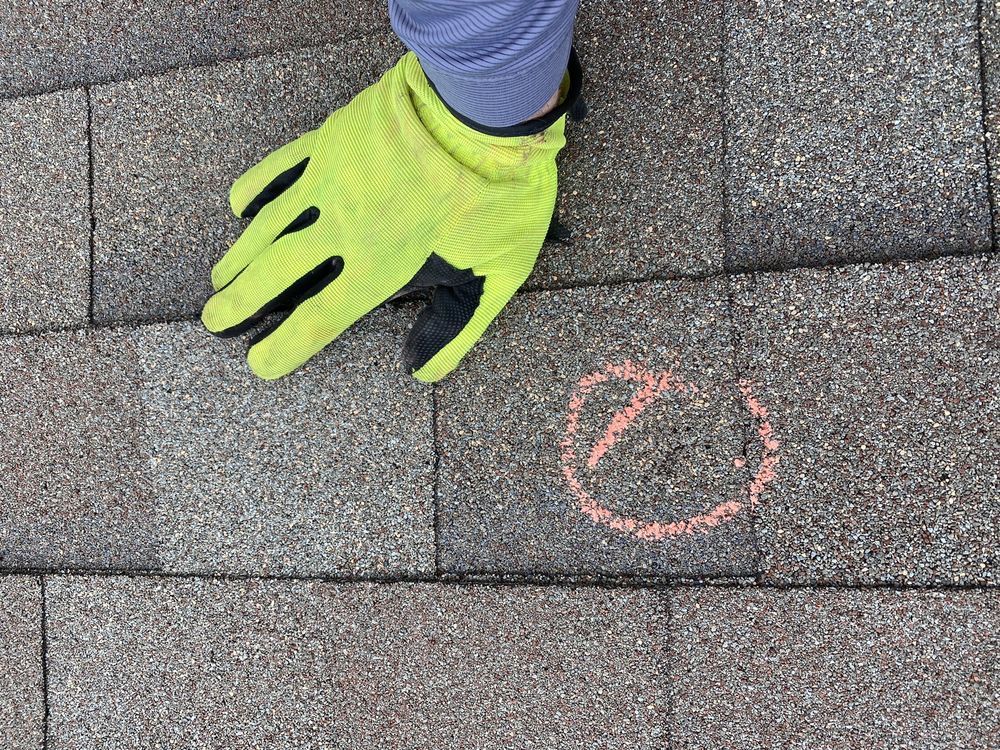 Gloved hand on a weathered shingle roof, marking a damaged spot with pink chalk.