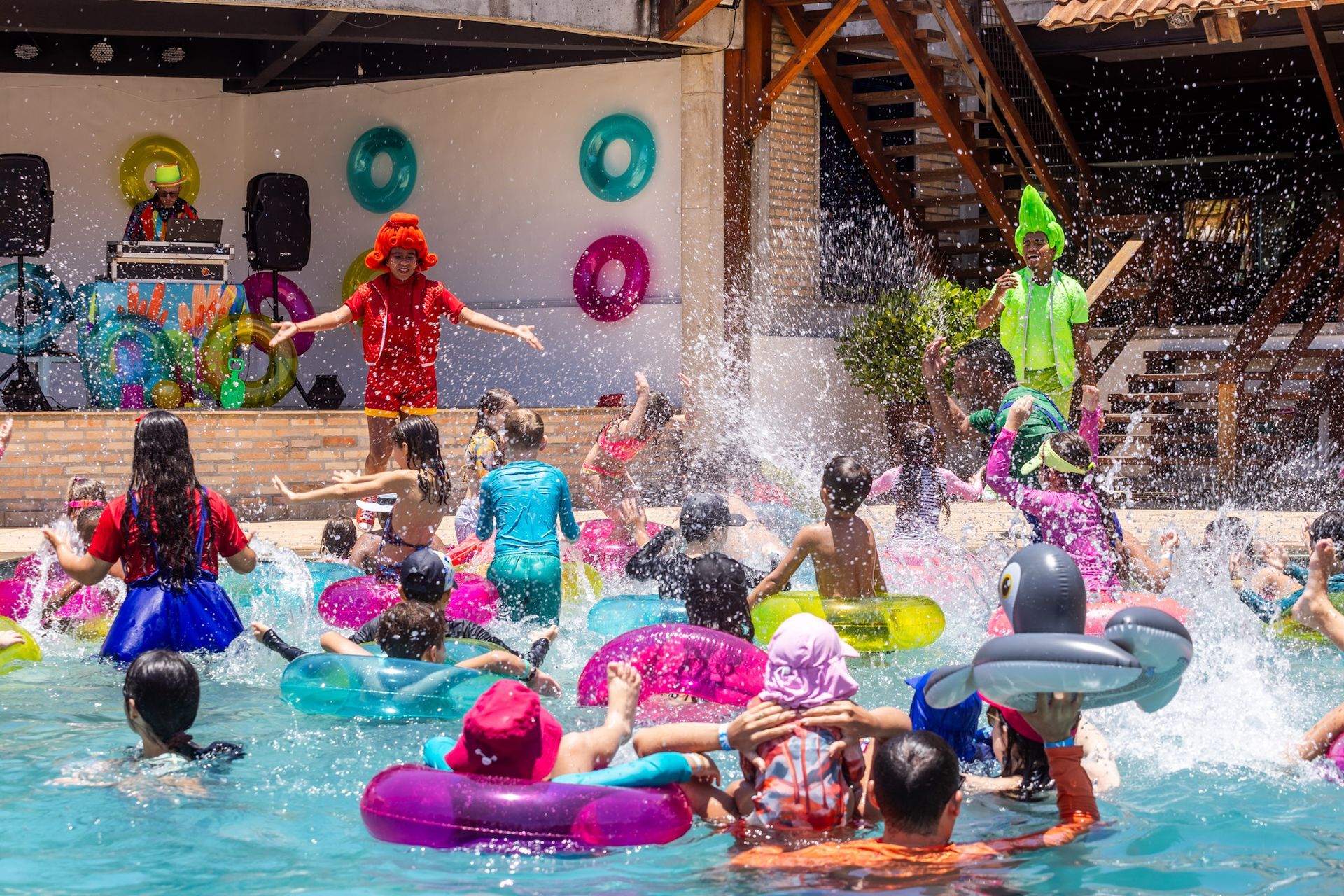 Um grupo de crianças na piscina, usando boias coloridas, participando de uma atividade animada. Dois animadores fantasiados de vermelho  e amarelo lideram a diversão na borda. Ao fundo, há um DJ com equipamentos de som e decoração temática com boias penduradas.