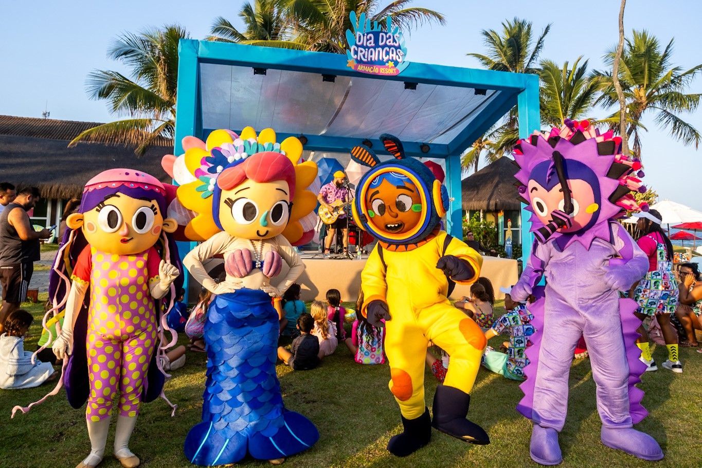 Os quatro personagens do Armação Resort (Cora, Pérola, Lui e Fluffy) estão em pé no gramado ao ar livre. Atrás deles tem um palco colorido com um show de música da banda Ludicanto. Muitas crianças estão sentadas no gramado assistindo o show. O dia está ensolarado, é fim de tarde e também existem palmeiras atrás do palco.