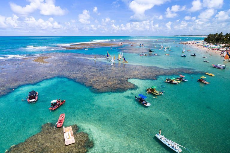 Vista aérea das piscinas naturais de Porto de Galinhas. Com vários barquinhos parados e jangadas. É possível ver a praia ao canto, com guarda-sóis e coqueiros.