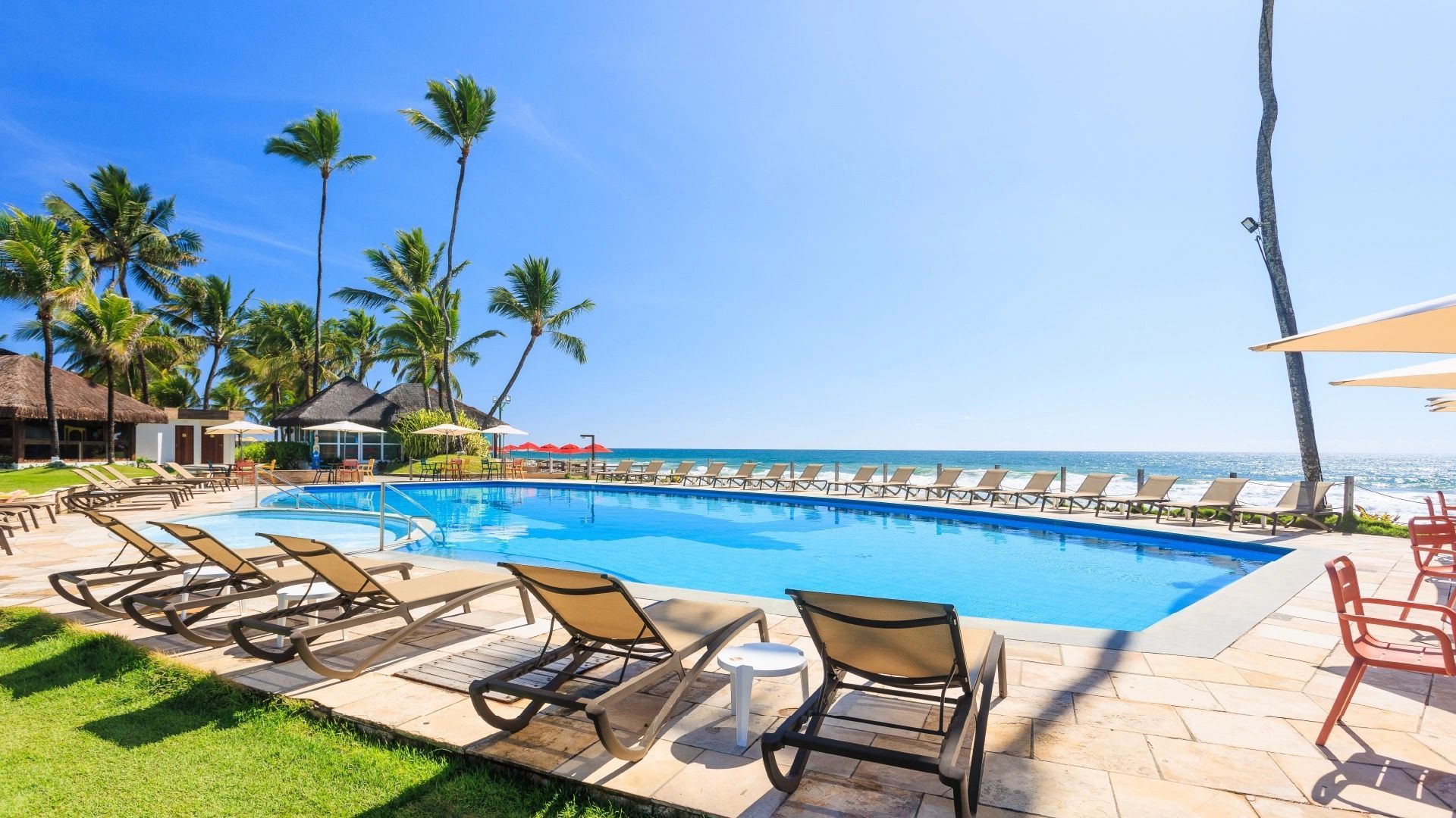 Uma piscina cercada por espreguiçadeiras e palmeiras, com vista para o mar. Céu azul e dia de sol.