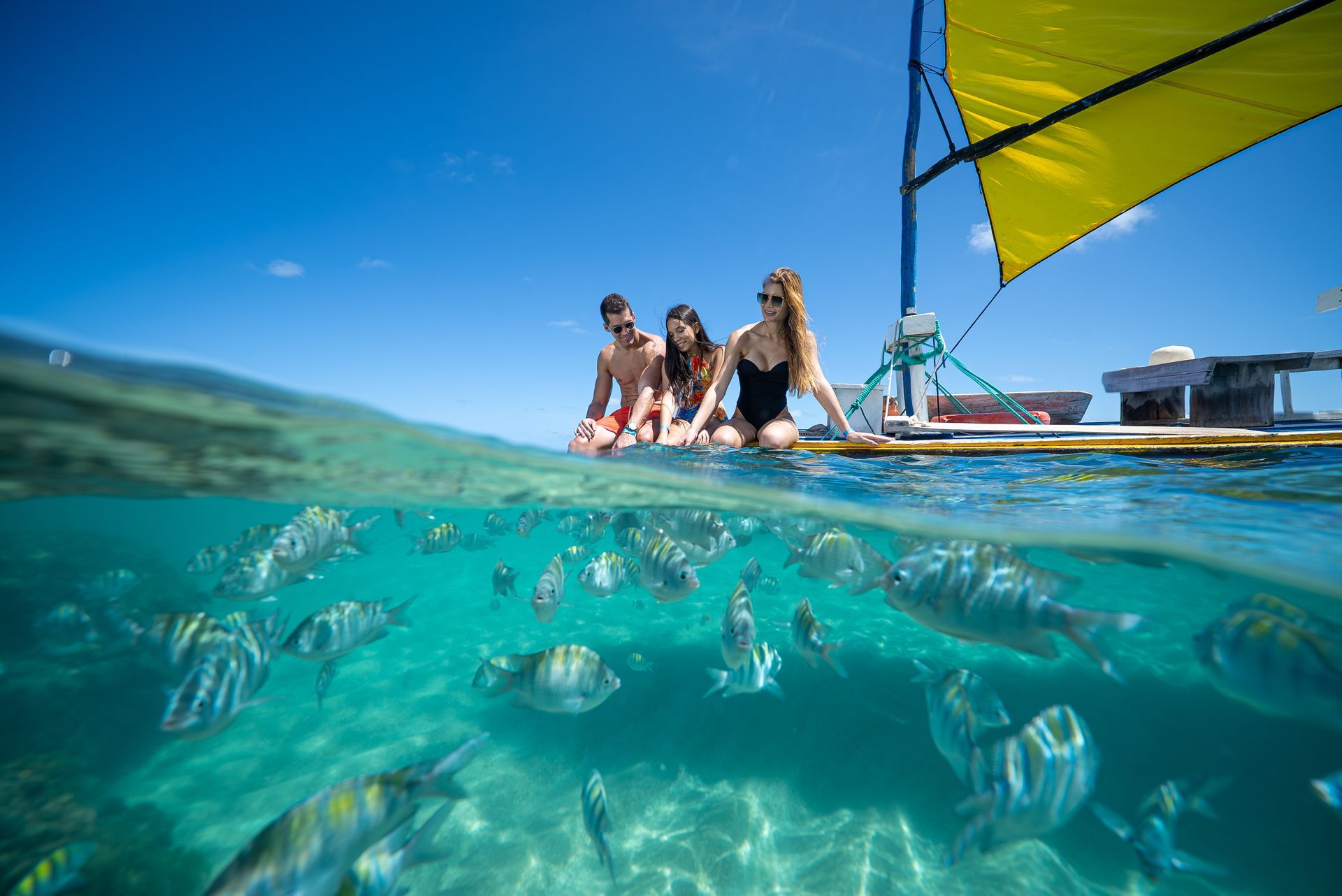 casal sentado em cadeiras sob guarda-sol na areia da praia, com mesa à frente com cocos e petiscos, enquanto duas crianças correm em direção ao mar com ondas ao fundo