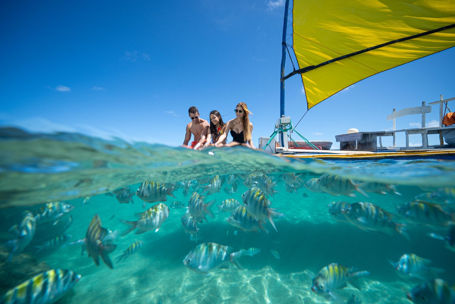 casal reunido à mesa em restaurante do resorts com janelas e vista para área verde, brindando com sucos e pratos servidos enquanto um bebê observa sentado ao lado na cadeirinha.