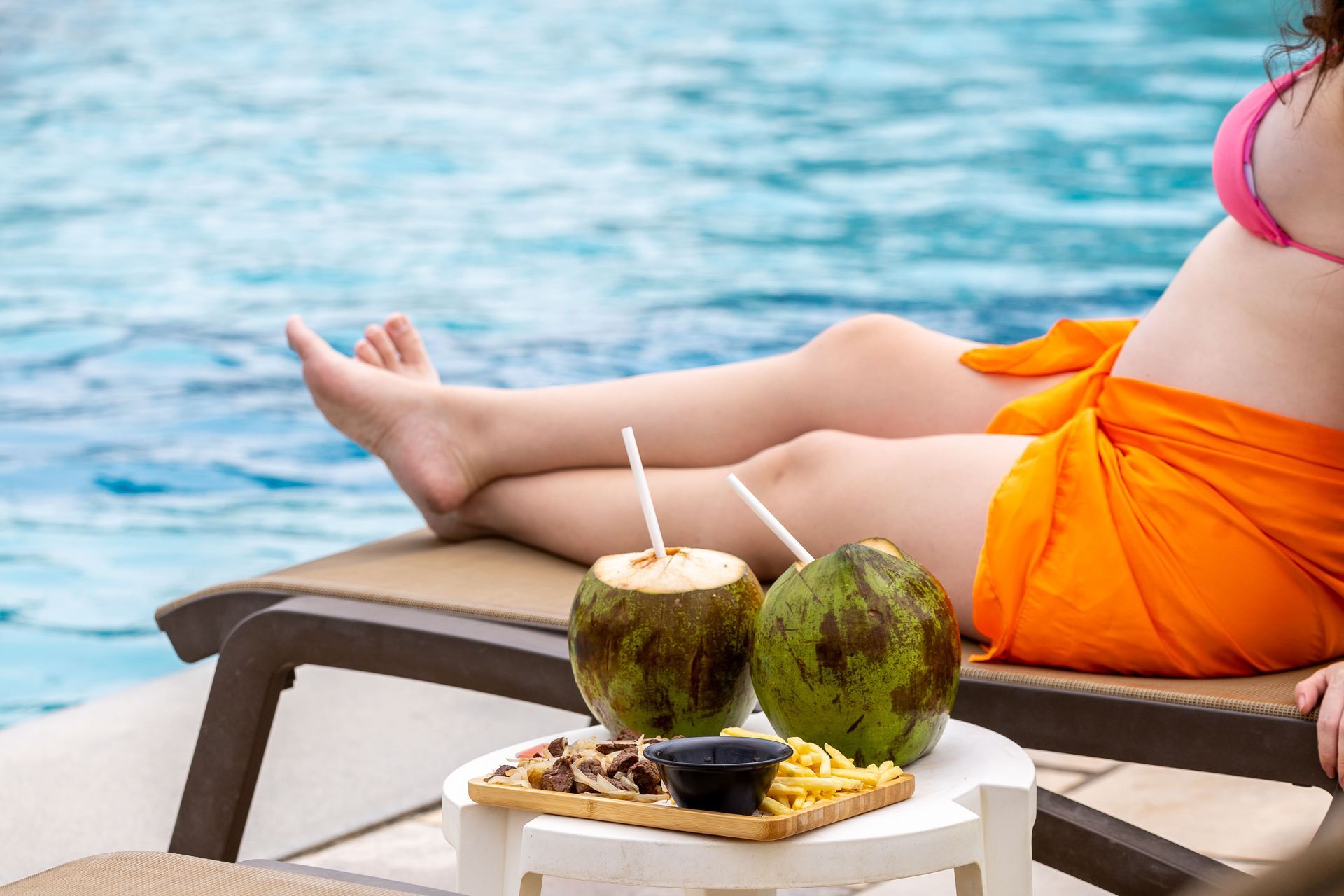 Mulher relaxando em uma espreguiçadeira à beira de uma piscina, com uma bandeja ao lado contendo cocos verdes e um petisco de carne com batata frita.