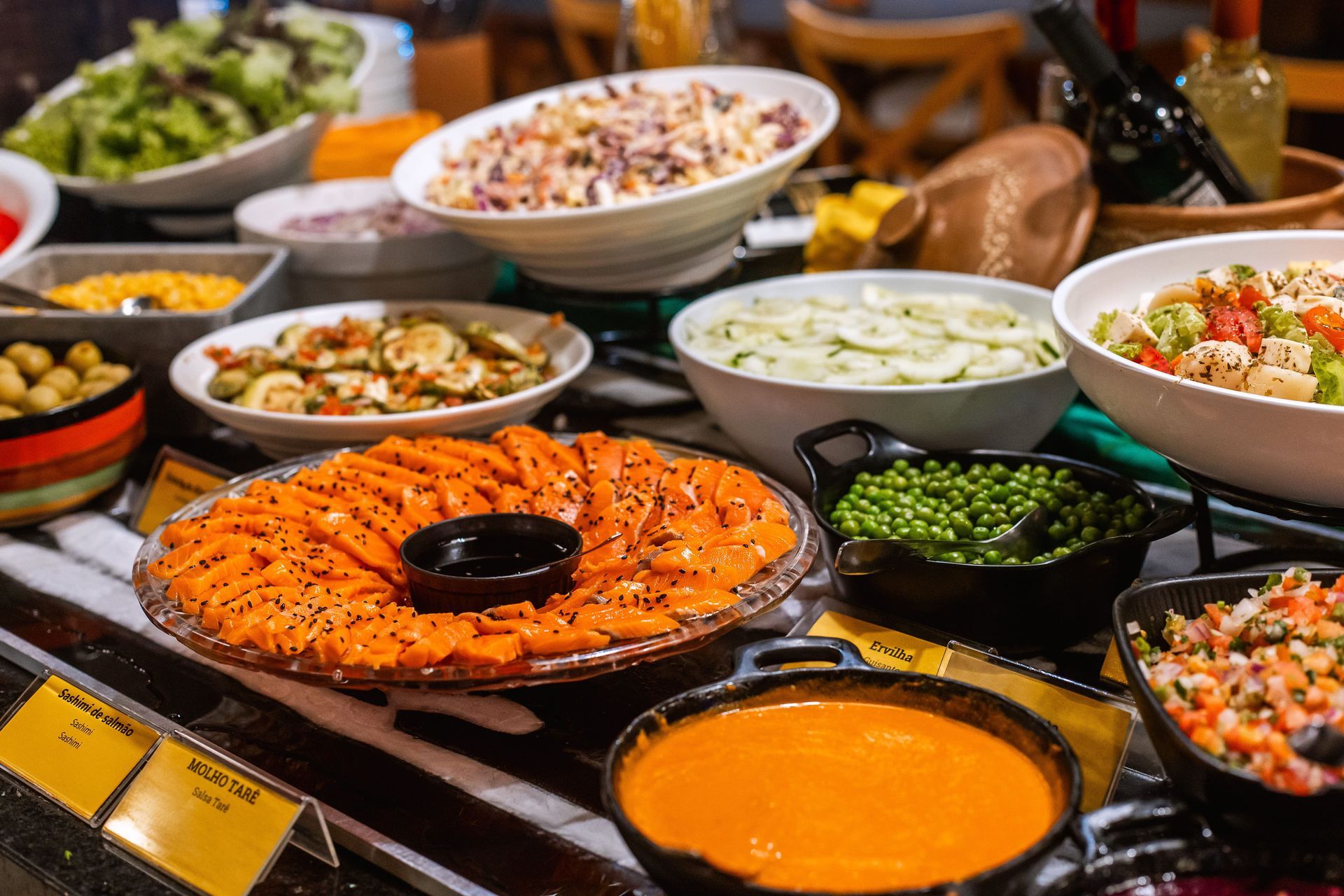 Buffet variado com diversas opções de saladas e acompanhamentos. Em destaque, um prato de salmão fatiado com gergelim e molho tare ao centro. Ao redor, há saladas de pepino, grão-de-bico, maionese, ervilhas, pimentões, vinagrete e folhas verdes. O ambiente tem iluminação aconchegante e decoração rústica.