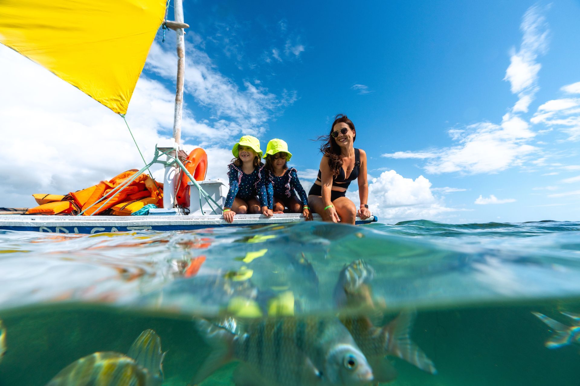 Mãe e duas filhas gêmeas, crianças, sentadas em jangada, nas piscinas naturais. É possível ver peixinhos embaixo da água cristalina. O dia está ensolarado e o céu azul.