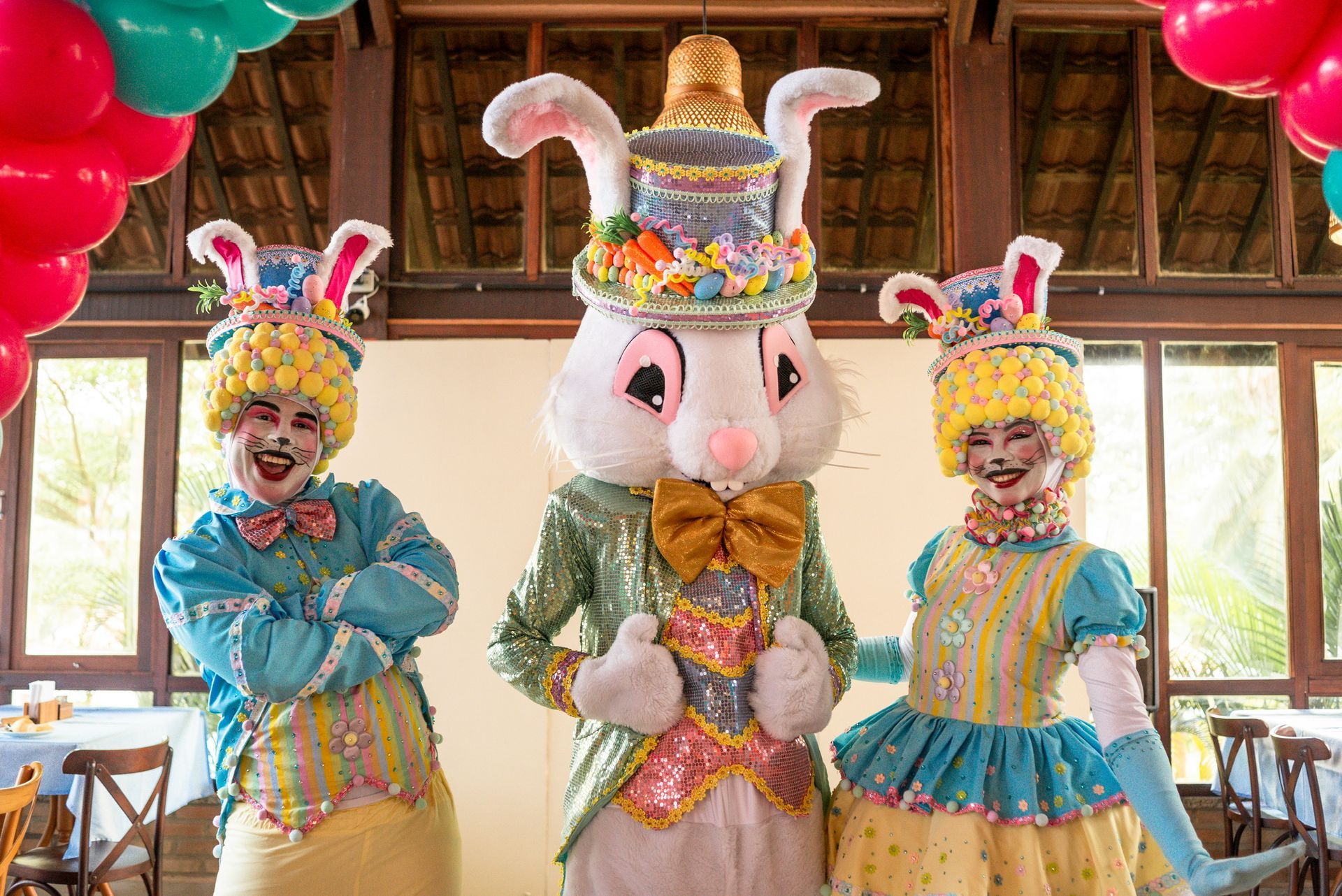 três personagens fantasiados de coelho da Páscoa com roupas coloridas e detalhes festivos posando em restaurante do resort decorado com balões e elementos temáticos