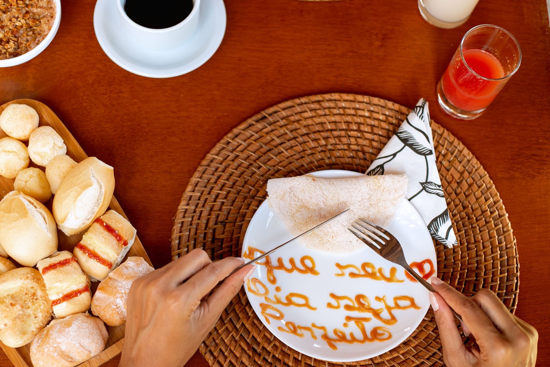 Pessoa cortando uma tapioca em um prato com a mensagem escrita em calda 'Que seu dia seja perfeito', acompanhada de pães variados, xícara de café e copo de suco.