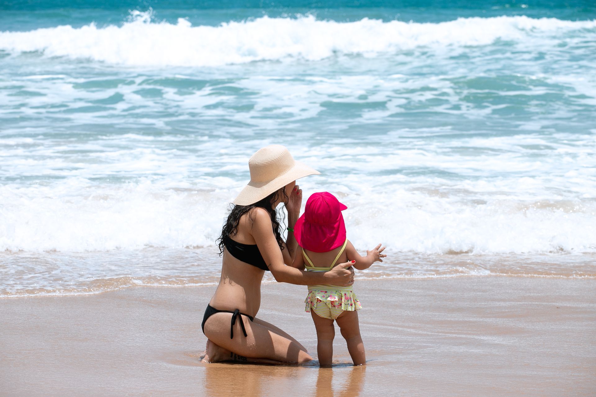 Mãe e filha bebê sentadas na areia da praia, observando as ondas do mar. As duas estão protegidas com chapéu e estão de frente para o mar, de costas para a câmera.