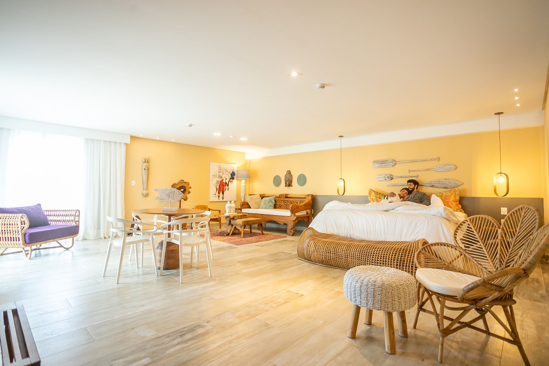 Quarto amplo e iluminado com decoração em tons claros, cama de casal, móveis de vime e madeira, mesa de jantar pequena e varanda com cortinas brancas.