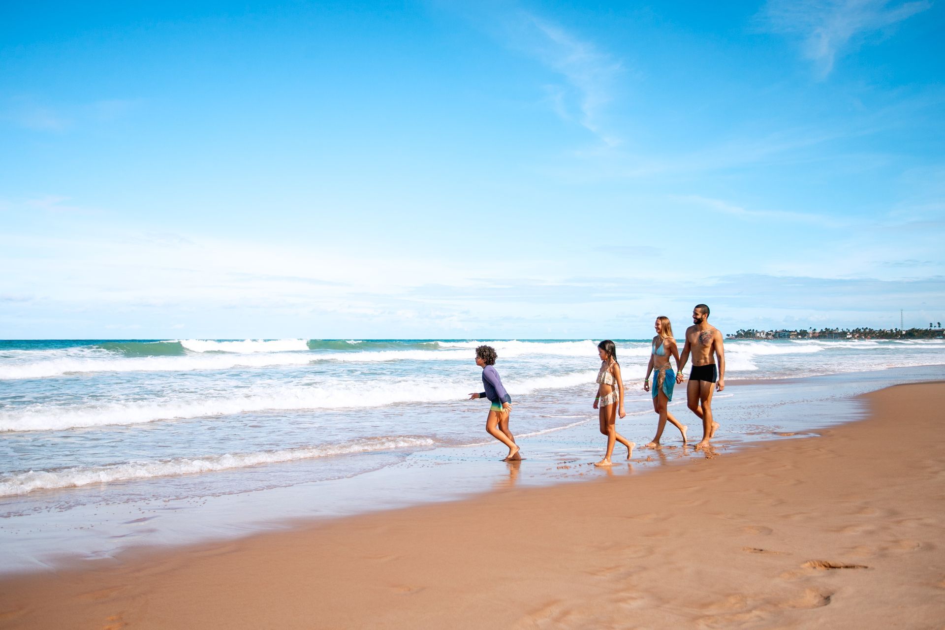 Família com pai, mãe e duas crianças (menino e menina), caminham na areia da praia de Porto de Galinhas. Ao fundo é possível ver o mar terminando em ondas na areia.