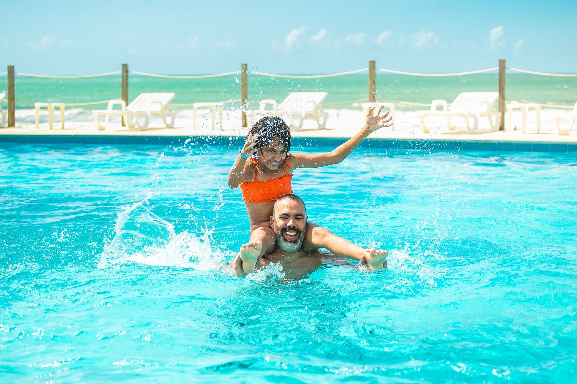 Pai e filha criança bricando na piscina. Ele tem a filha nos ombros e os dois estão sorrindo. Ao fundo é possível ver a praia com mar verde.