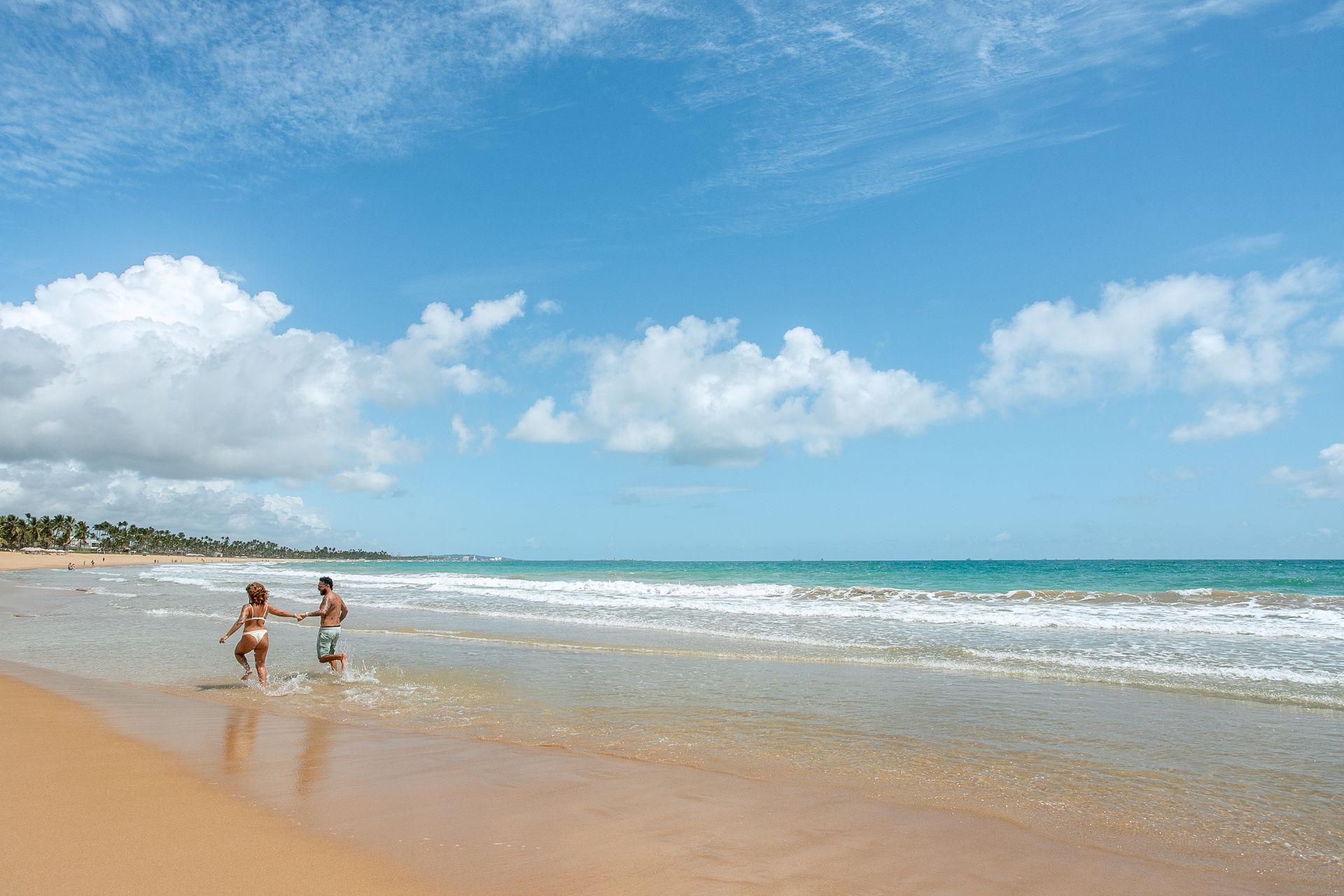 Praia do Cupe. Um casal de homem e mulher correm à beira-mar, o dia está ensolarado e o céu azul. Atrás deles está um mar azul e ao fundo é possível ver palmeiras.