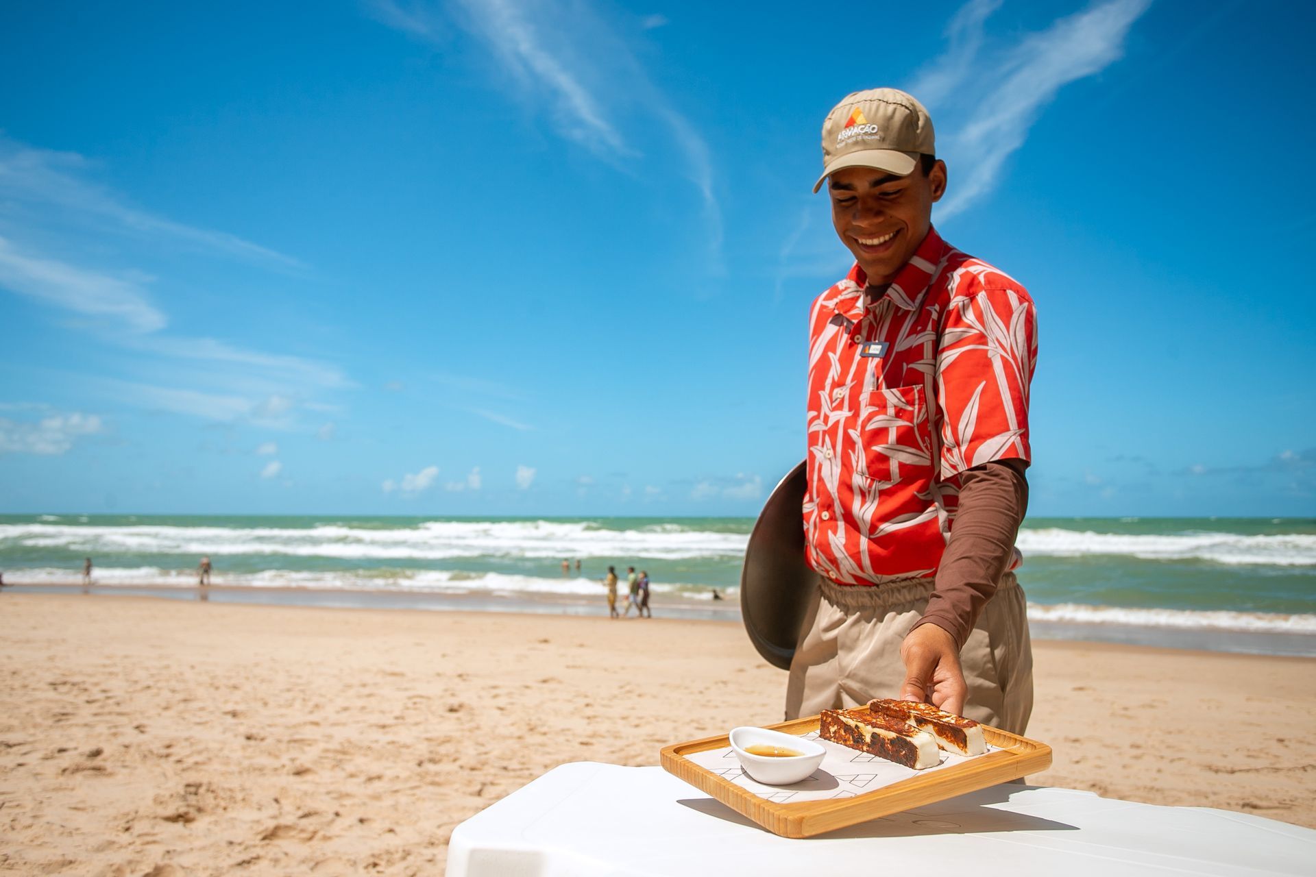 Garçom sorridente vestindo camisa vermelha com estampa tropical, boné bege e calça clara, servindo uma bandeja com queijo coalho e mel em uma mesa na areia da praia. Ao fundo, o mar com ondas, céu azul com poucas nuvens e algumas pessoas caminhando na beira da água, criando uma atmosfera alegre e acolhedora à beira-mar.