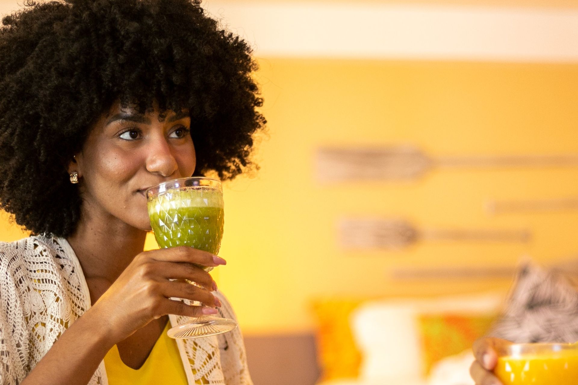 Mulher bebendo suco verde em taça de vidro, dentro do quarto do resort.