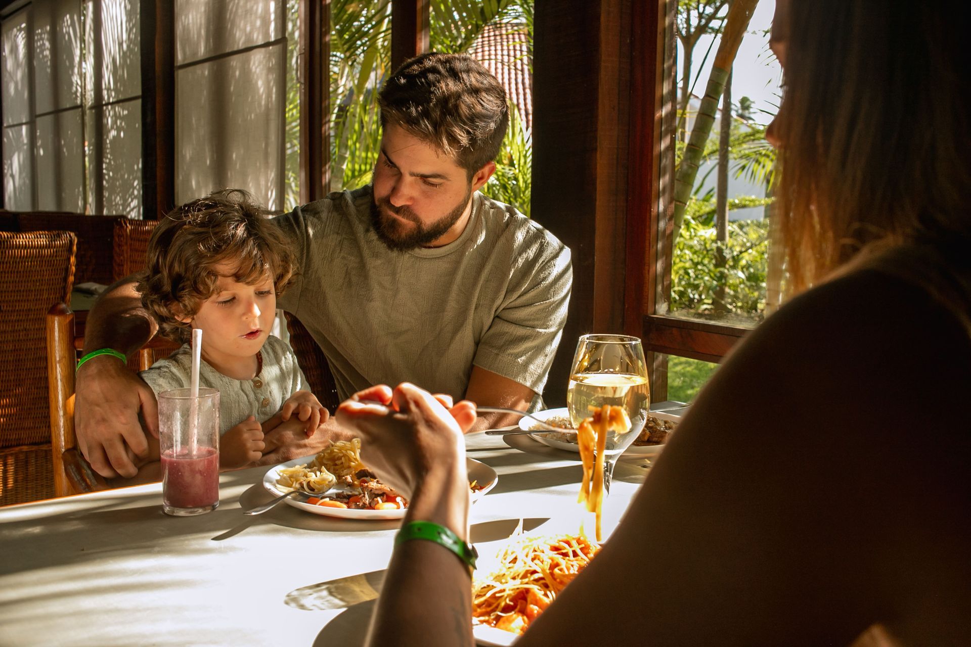 Família reunida em um restaurante iluminado pela luz natural. Um homem ajuda uma criança pequena a comer, enquanto uma mulher, de costas para a câmera, segura um garfo com macarrão. A mesa está posta com pratos de comida, uma taça de vinho branco e um suco de frutas. Ao fundo, grandes janelas de madeira revelam uma paisagem verde com árvores e plantas tropicais.