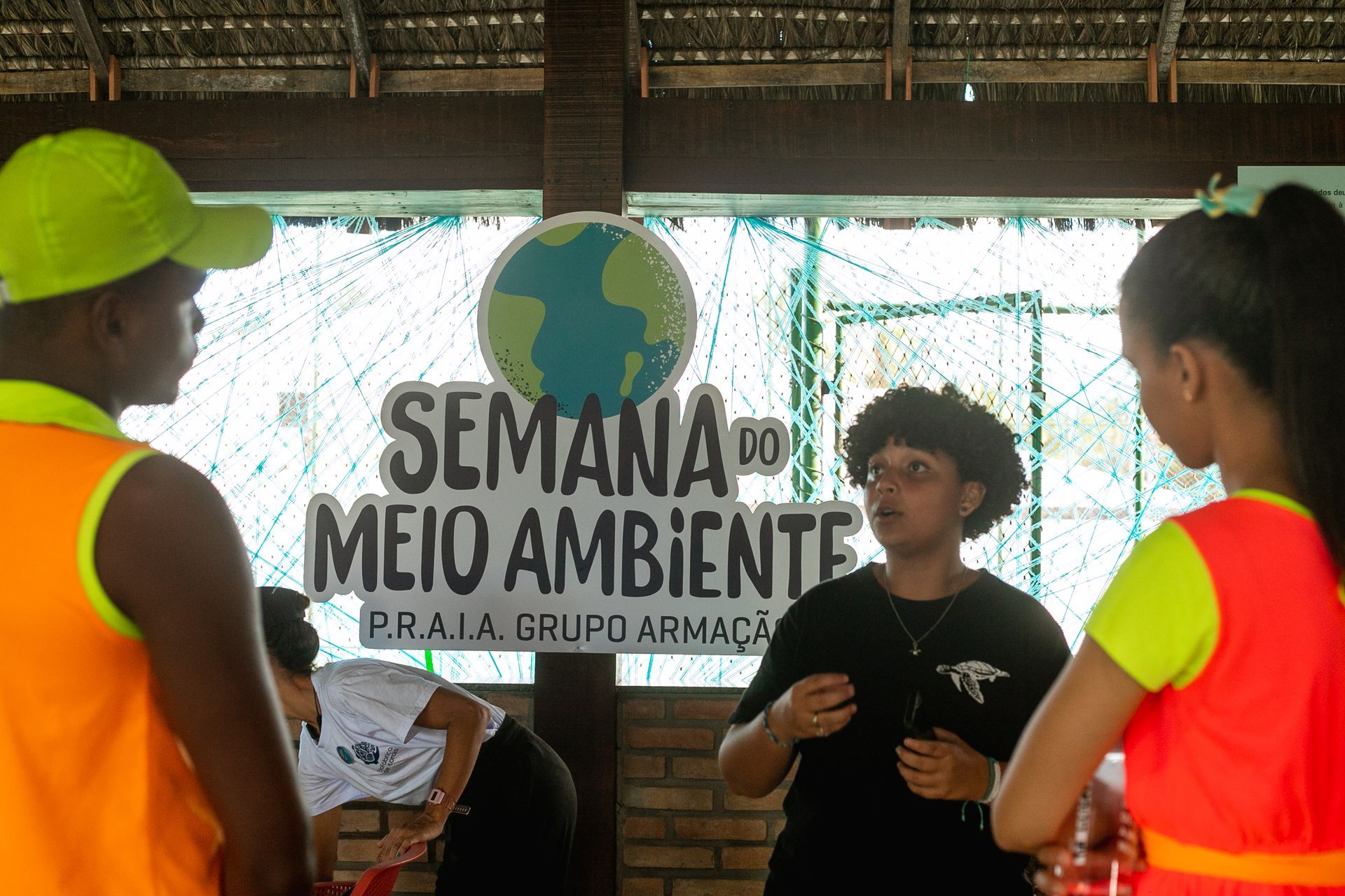 Pessoas reunidas em atividade educativa da Semana do Meio Ambiente promovida pelo Grupo Armação, com um cartaz ao fundo destacando o nome do evento.