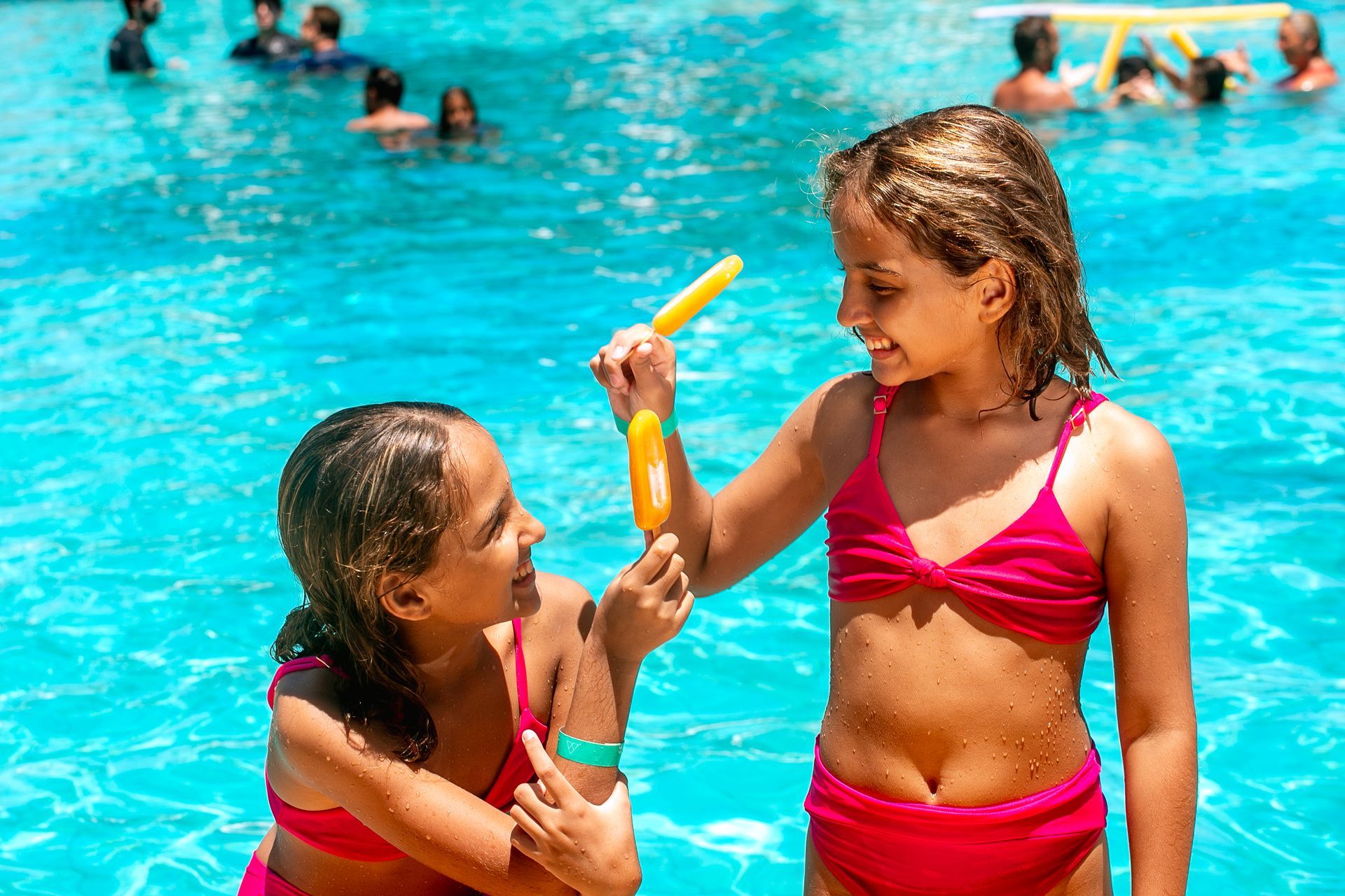 Duas meninas de biquini rosa seguram picolés. A piscina azul aparece ao fundo.