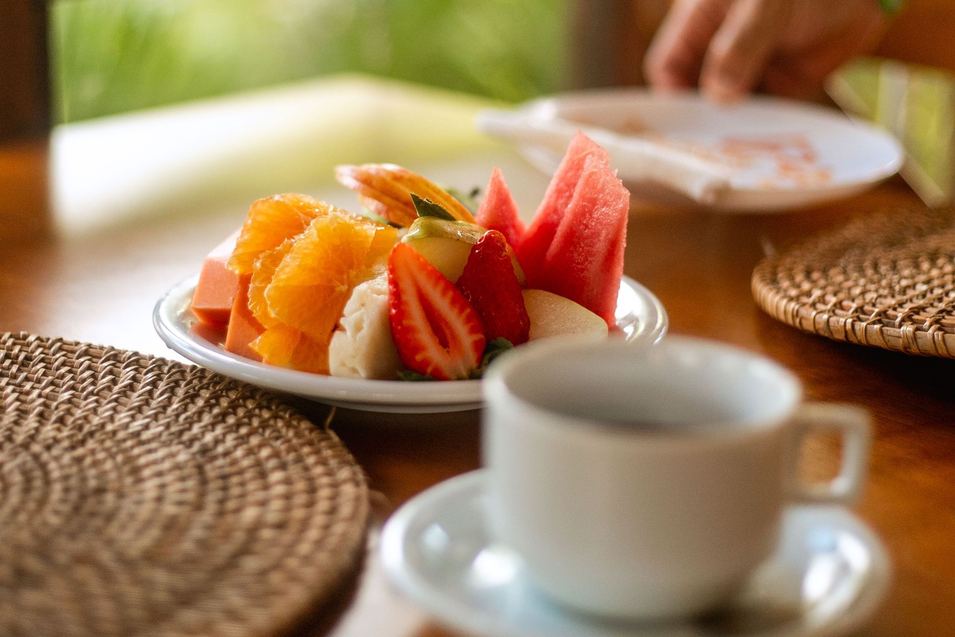 Prato com frutas frescas, incluindo fatias de laranja, morango, melancia e mamão, acompanhado por uma xícara de café sobre uma mesa com lugar americano de palha.