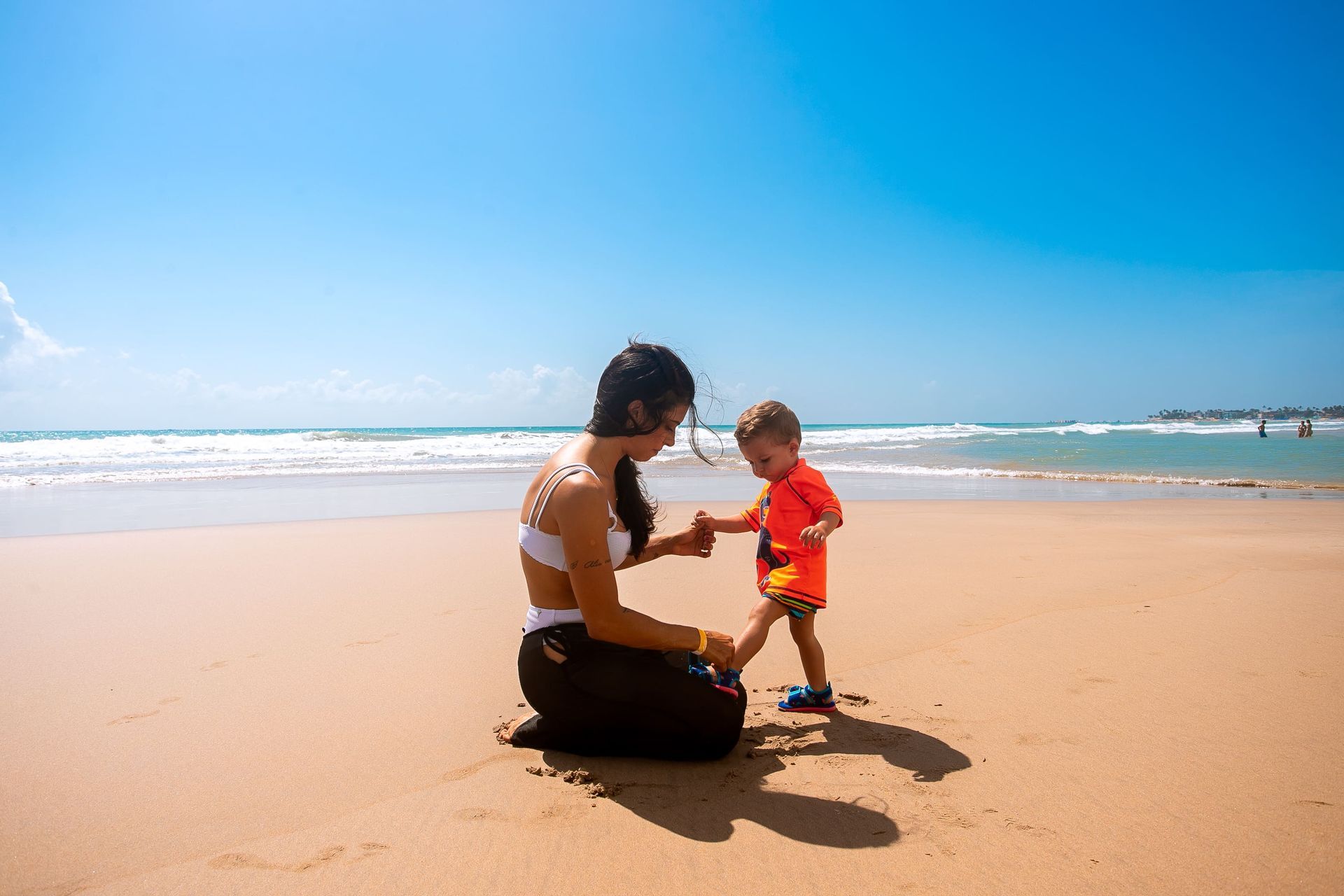 Uma mulher ajoelhada na areia segura a mão de uma criança pequena que dá os primeiros passos na praia. O mar e o céu azul compõem o cenário ao fundo.