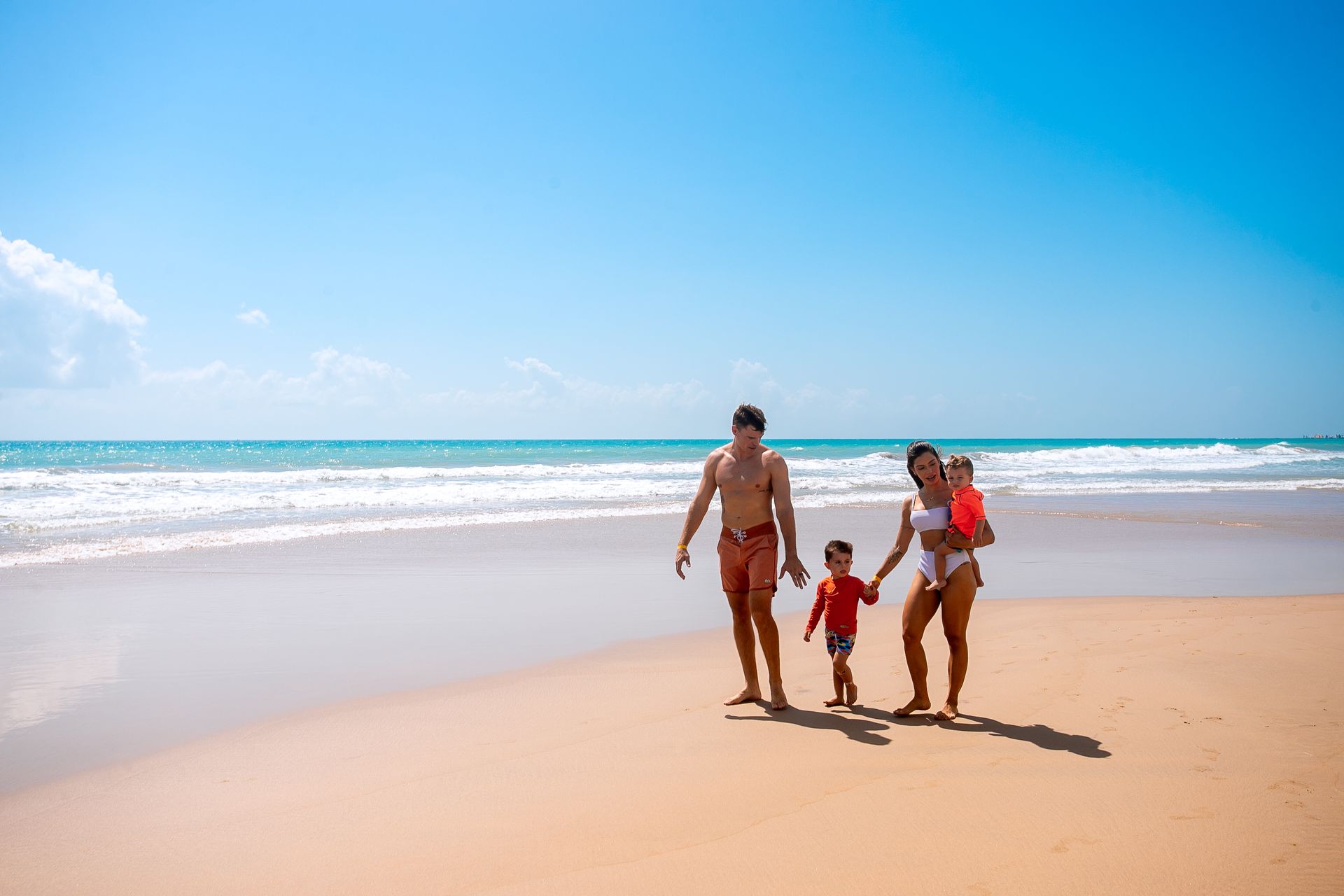 Família com pai, mãe e dois meninos pequenos caminhando na praia. O céu e o mar estão azuis e o dia ensolarado. A maré está baixa e a areia lisinha.