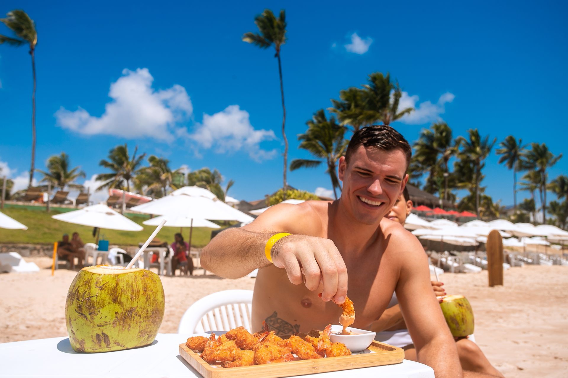 Homem aproveitando um dia ensolarado na praia, comendo petiscos de camarão empanado e bebendo água de coco, com guarda-sóis e coqueiros ao fundo.