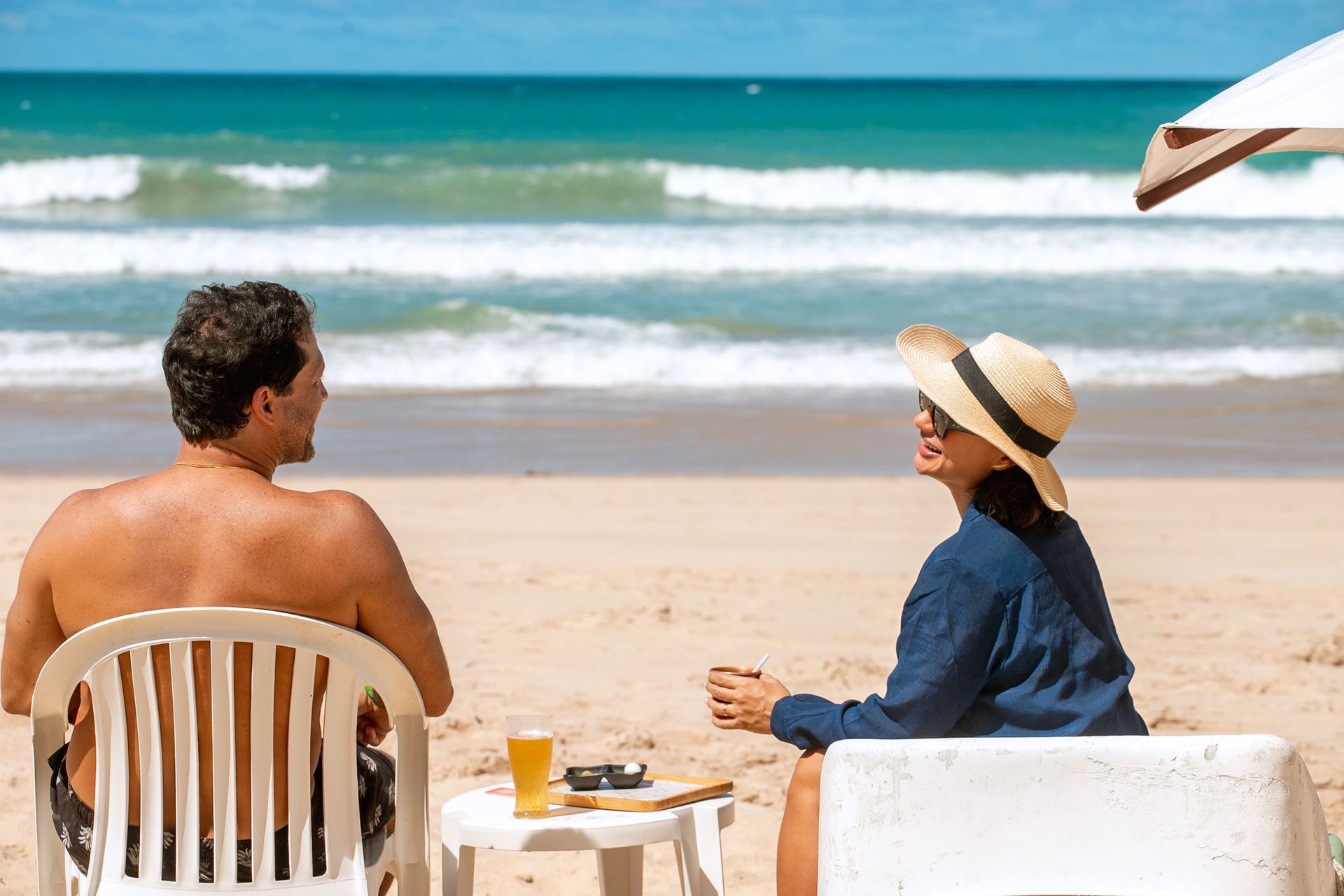 Homem e mulher sentados em cadeiras na praia, com vista para o mar verde. O dia está ensolarado.