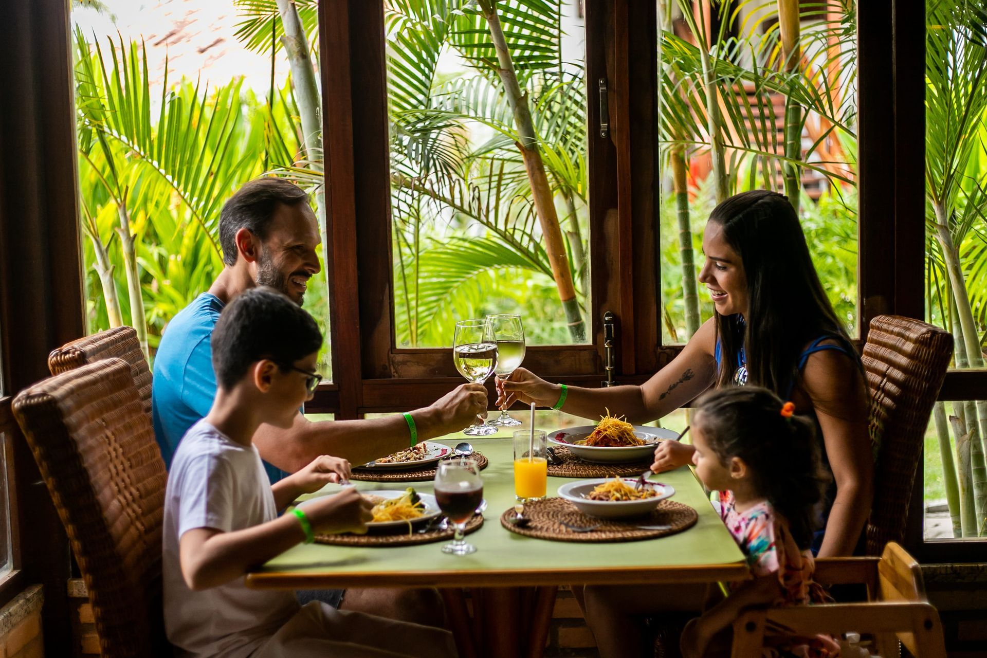 Família reunida em um restaurante aconchegante durante o dia, desfrutando de uma refeição juntos. O casal, sorridente, brinda com taças de vinho branco, enquanto as crianças comem macarrão. A mesa está decorada com jogos americanos de palha e bebidas variadas, incluindo suco de laranja e refrigerante.