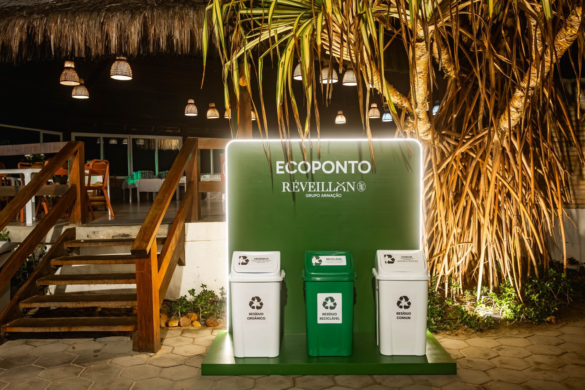 Estrutura de coleta seletiva chamada “Ecoponto Réveillon”, com três lixeiras identificadas para resíduos orgânicos, recicláveis e comuns, posicionadas em frente a um restaurante com decoração rústica. Ao redor há plantas tropicais, escada de madeira e iluminação quente pendente, criando um ambiente noturno acolhedor e sustentável.
