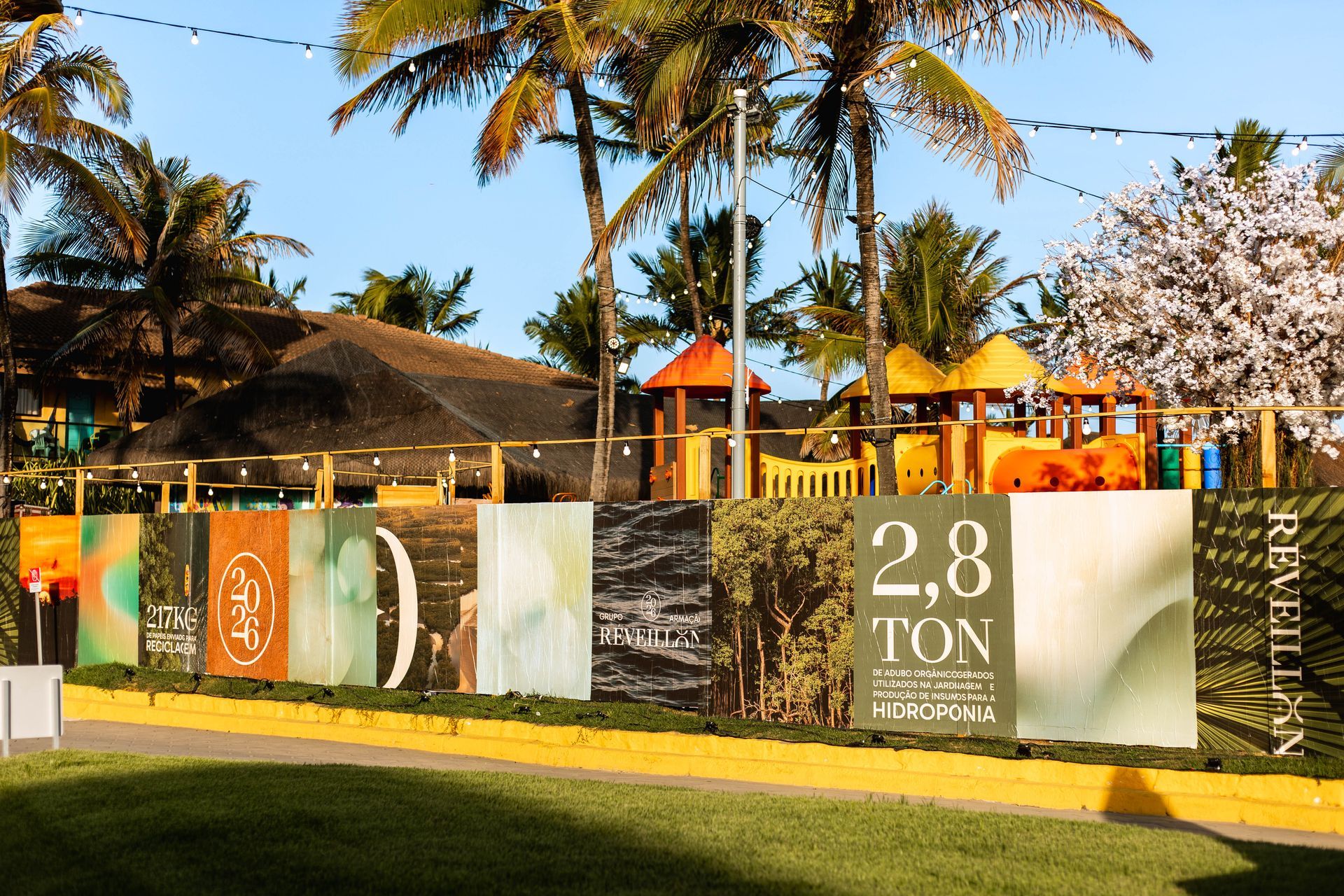 Painéis informativos ao ar livre exibem dados de sustentabilidade, em frente a uma área com palmeiras, playground colorido e estruturas de lazer. O cenário é tropical, com gramado, iluminação decorativa suspensa e construções ao fundo sob luz natural do fim de tarde.