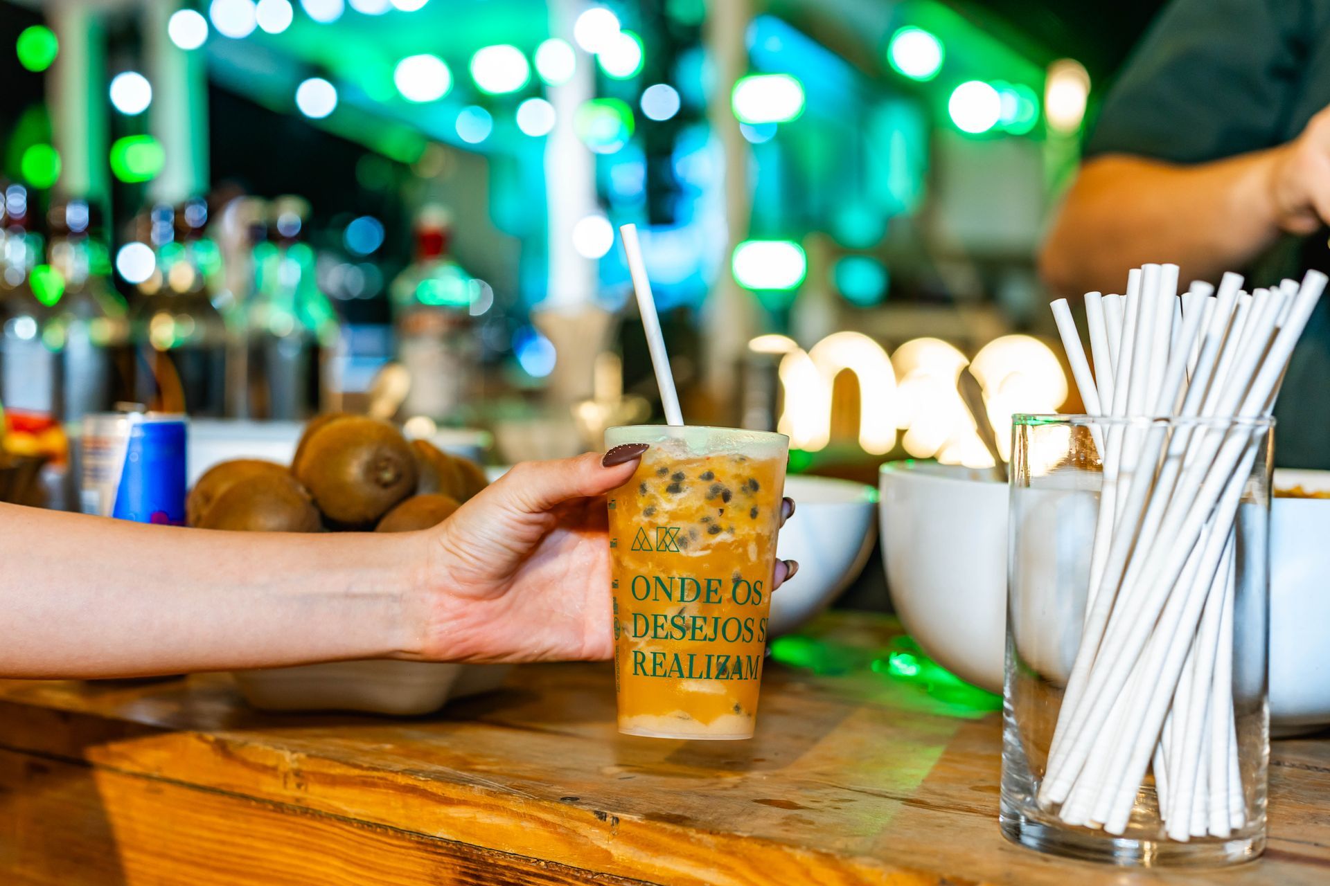 Close-up de uma mão segurando um copo de bebida. No copo está escrito “ONDE OS DESEJOS SE REALIZAM”. Ao redor, há um pote com canudos brancos, frutas e garrafas ao fundo, com iluminação verde desfocada que cria um clima moderno e noturno.
