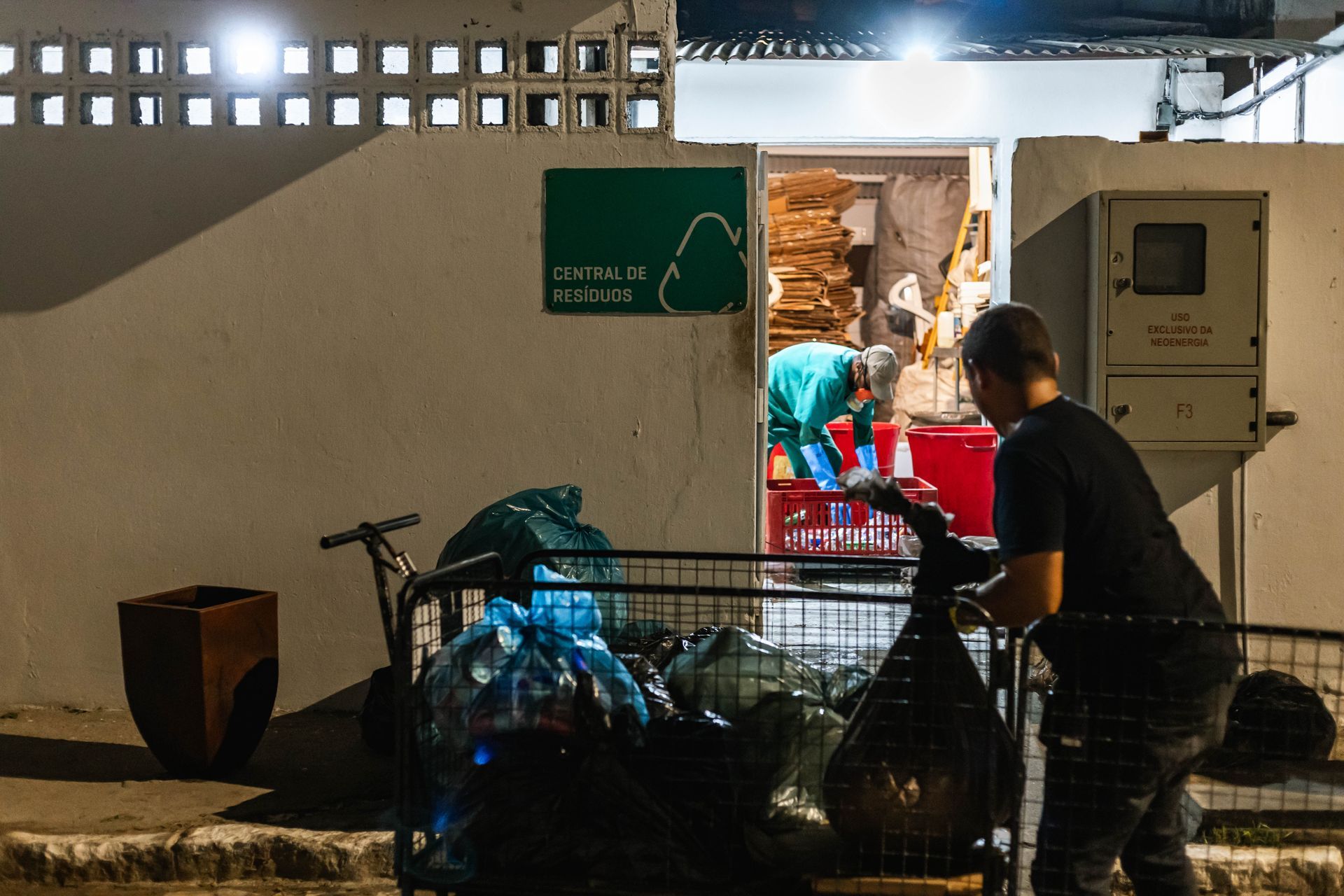 Área de serviço identificada como “Central de Resíduos”, onde trabalhadores fazem a separação e o manuseio de lixo reciclável. Em primeiro plano, há carrinhos cheios de sacos de resíduos; ao fundo, uma pessoa trabalha com caixas de papelão dentro do espaço iluminado, em um ambiente funcional e organizado.