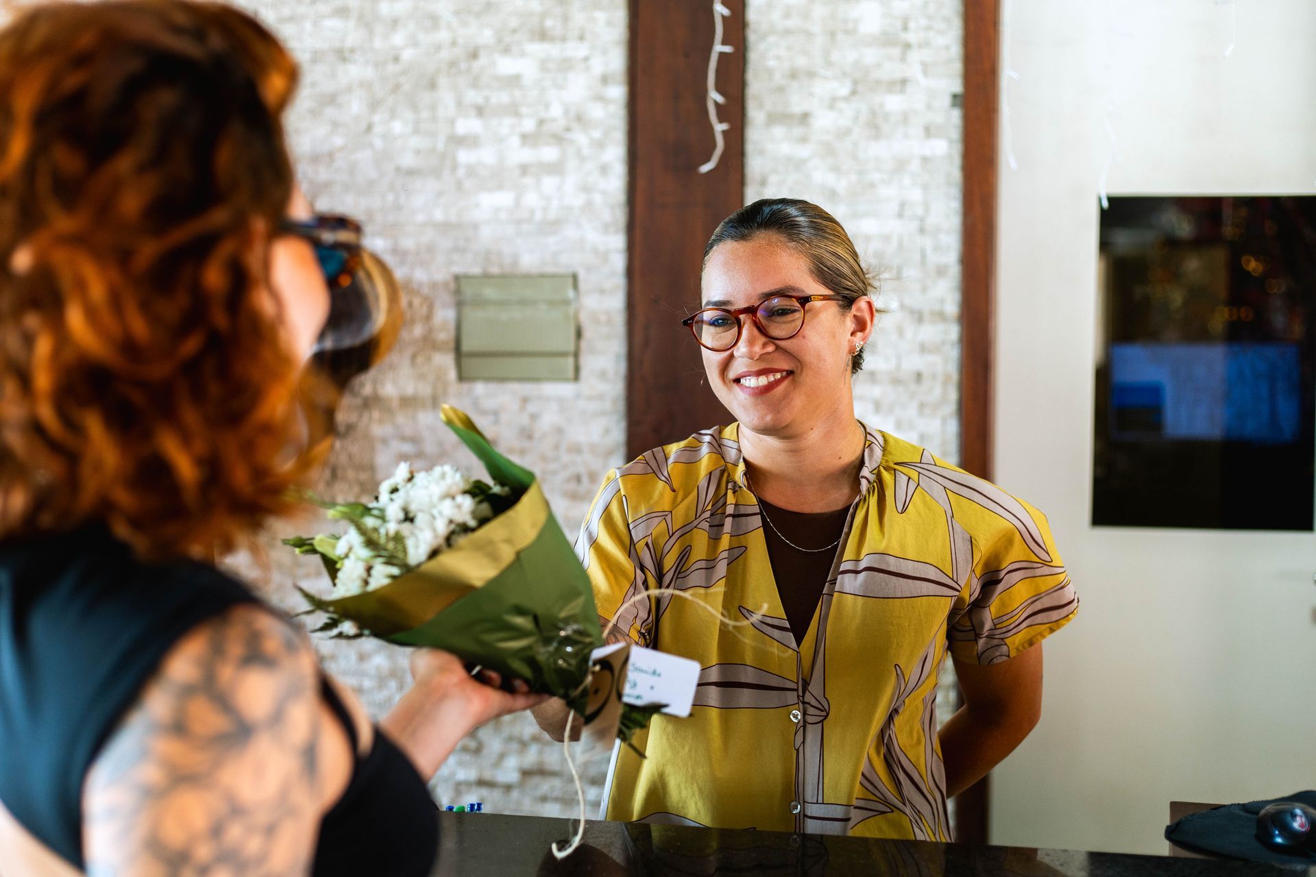 Mulher usando camisa amarela estampada e óculos entrega um pequeno buquê de flores a outra mulher em uma recepção de hotel. Ela sorri durante o atendimento atrás do balcão, em um ambiente interno com iluminação aconchegante, transmitindo acolhimento e cordialidade.