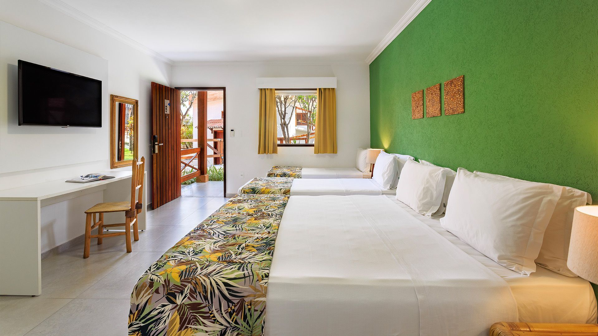 Quarto amplo com duas camas de casal, parede verde, roupas de cama com estampa tropical, mesa de trabalho com TV e porta de madeira aberta para varanda externa.