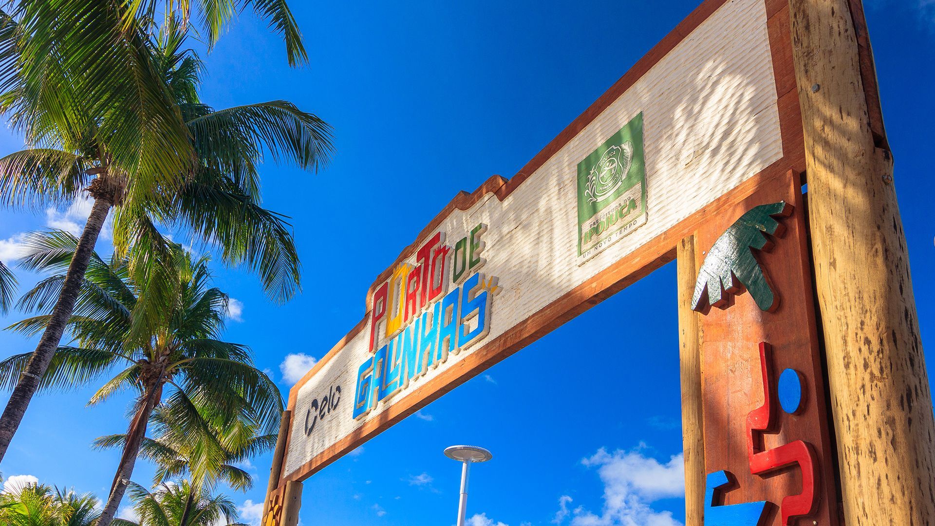 Placa colorida de entrada de Porto de Galinhas, cercada por coqueiros sob um céu azul claro.