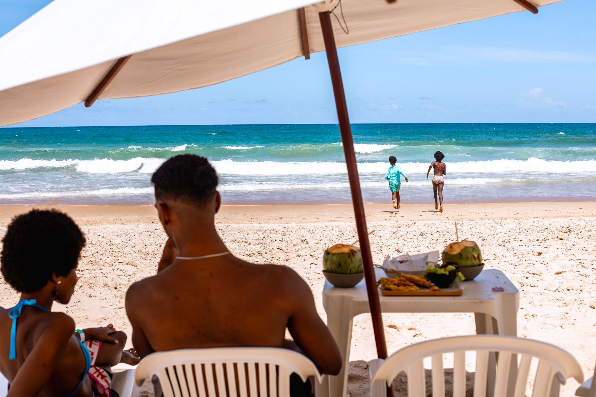 casal sentado em cadeiras sob guarda-sol na areia da praia, com mesa à frente com cocos e petiscos, enquanto duas crianças correm em direção ao mar com ondas ao fundo