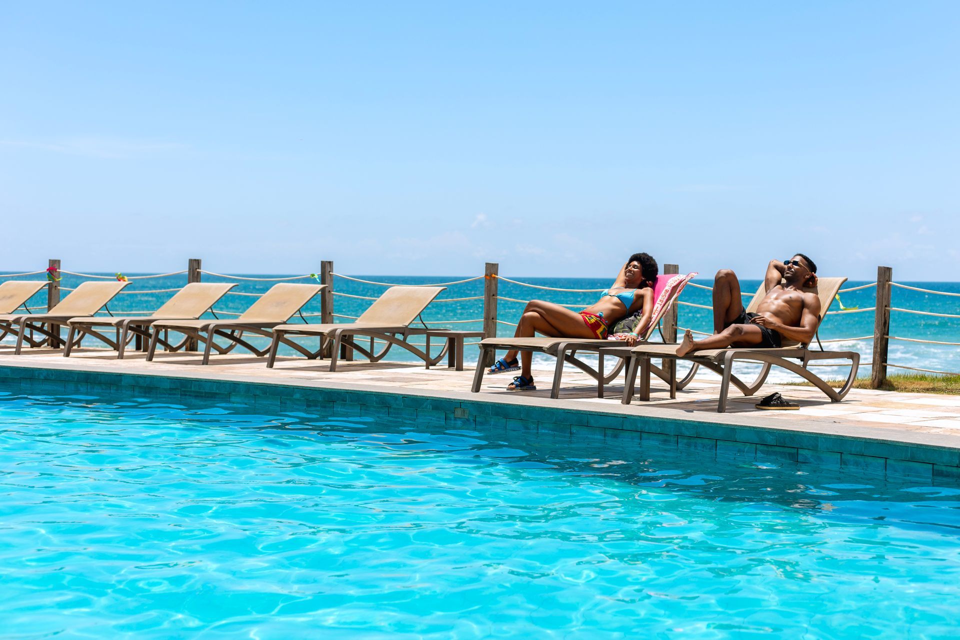 Casal relaxa em espreguiçadeiras à beira da piscina com vista para o mar azul, sob céu limpo e clima