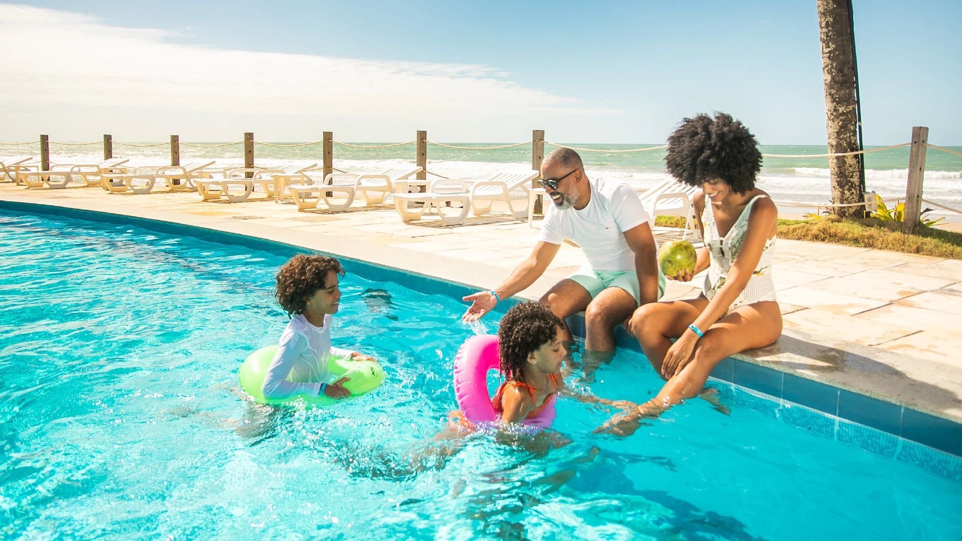 Família curtindo a piscina à beira-mar. Os pais estão sentados na borda e as duas crianças na água.