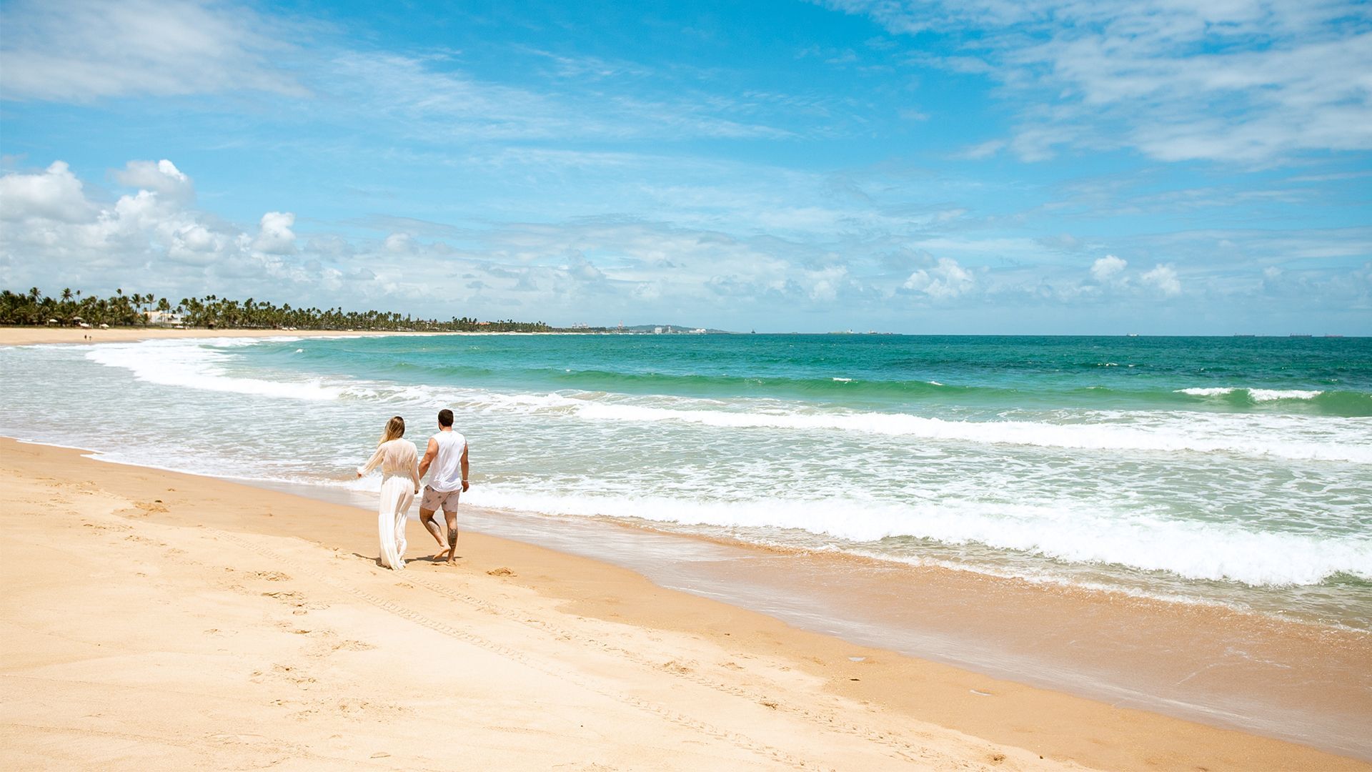 Casal caminhando na praia. O céu está azul, o mar está verde, é possível ver coqueiros ao longe.