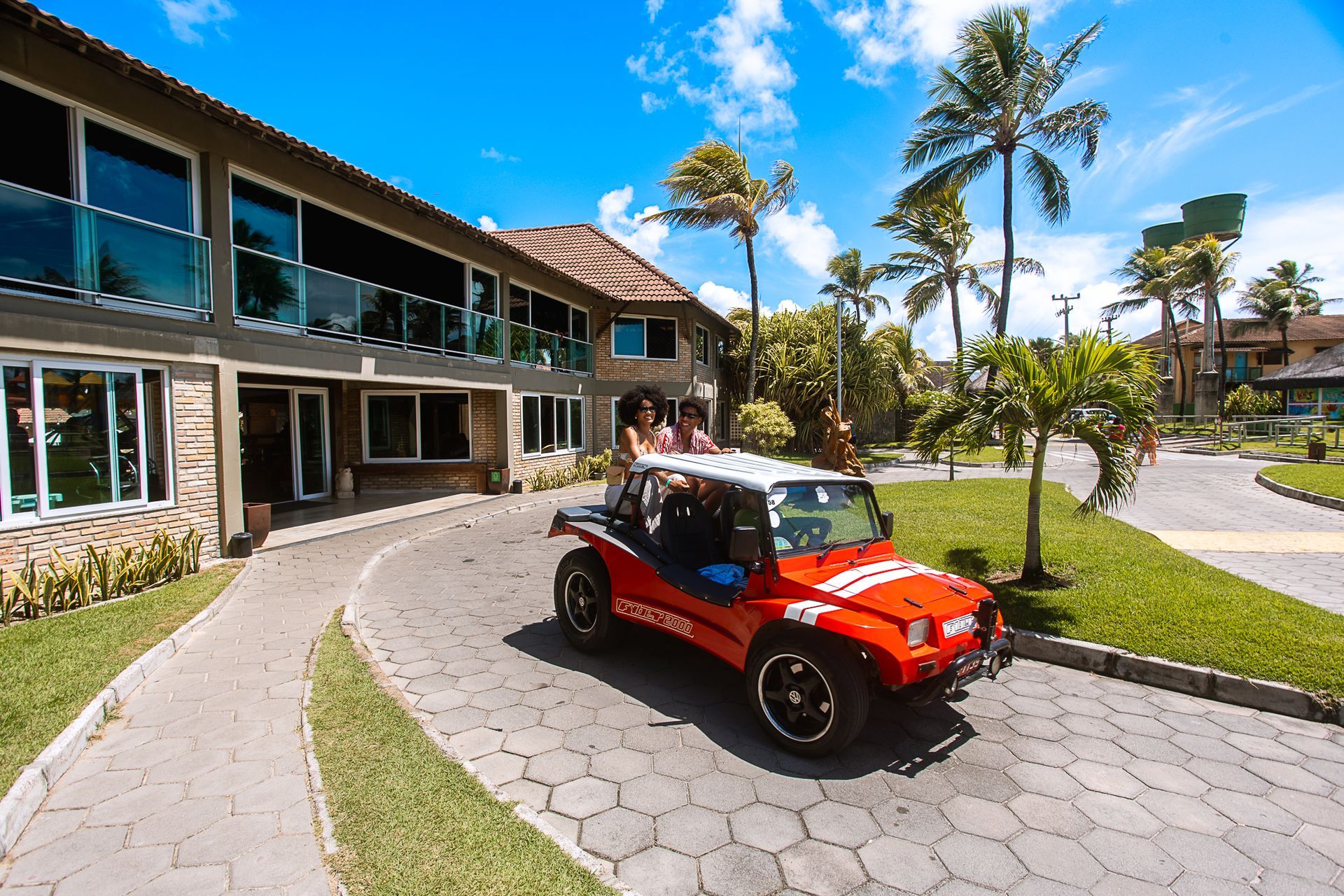 Hóspedes passeando de buggy vermelho pelo Armação Resort em Porto de Galinhas, com céu azul, palmeiras ao vento e estrutura do resort ao fundo.