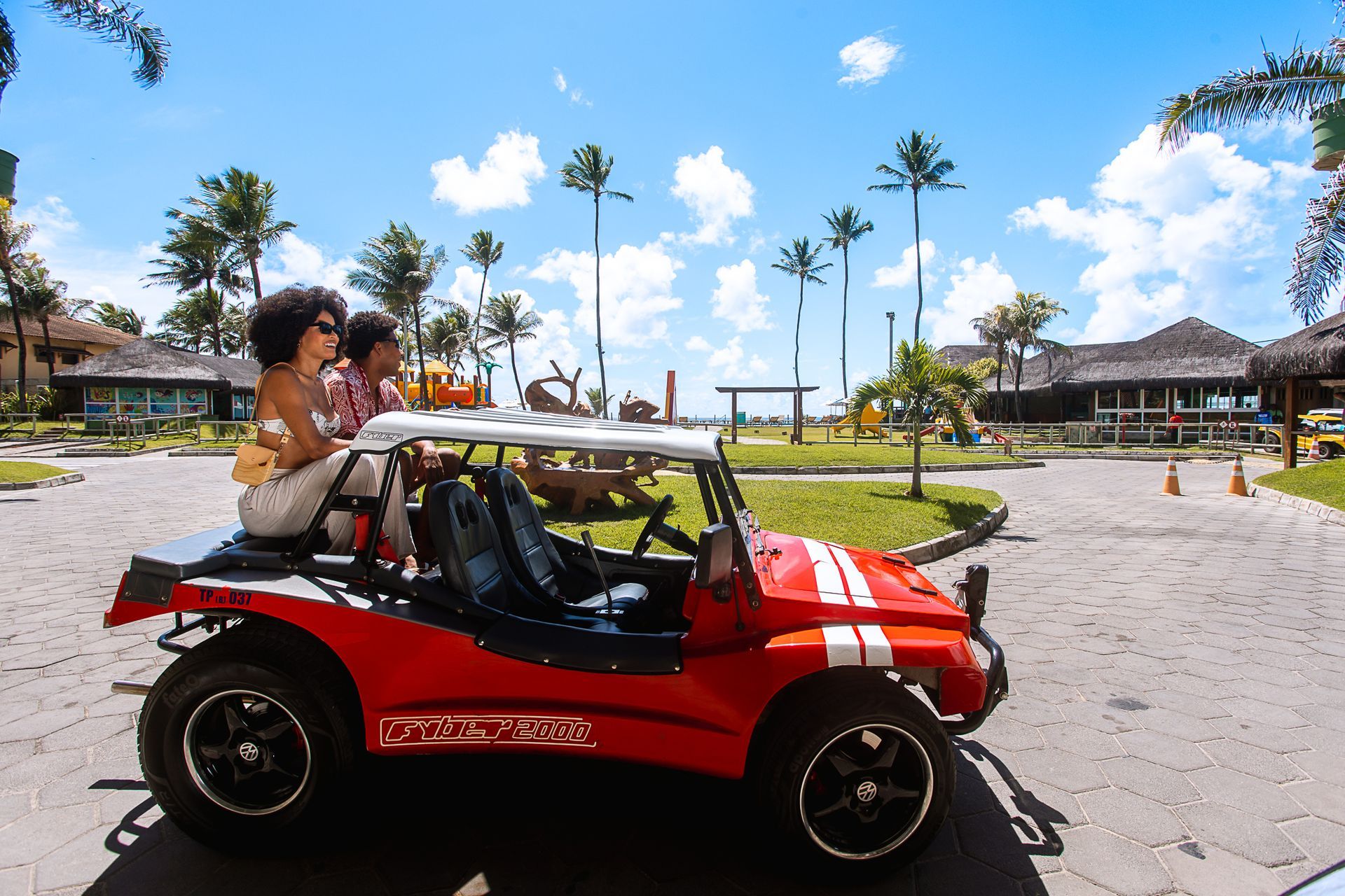 Duas pessoas sentadas em um buggy vermelho conversível estacionado em um resort tropical, com palmeiras, céu azul, playground ao fundo e construções com telhado de palha em um dia ensolarado.