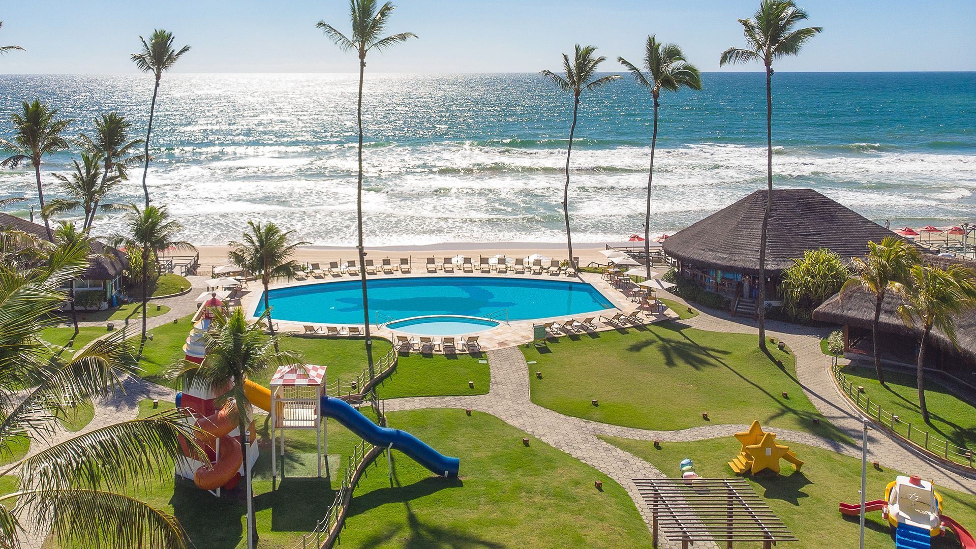 Vista aérea do Armação Resort. É possível ver praia, piscina, grama verde do resort, brinquedos playground.