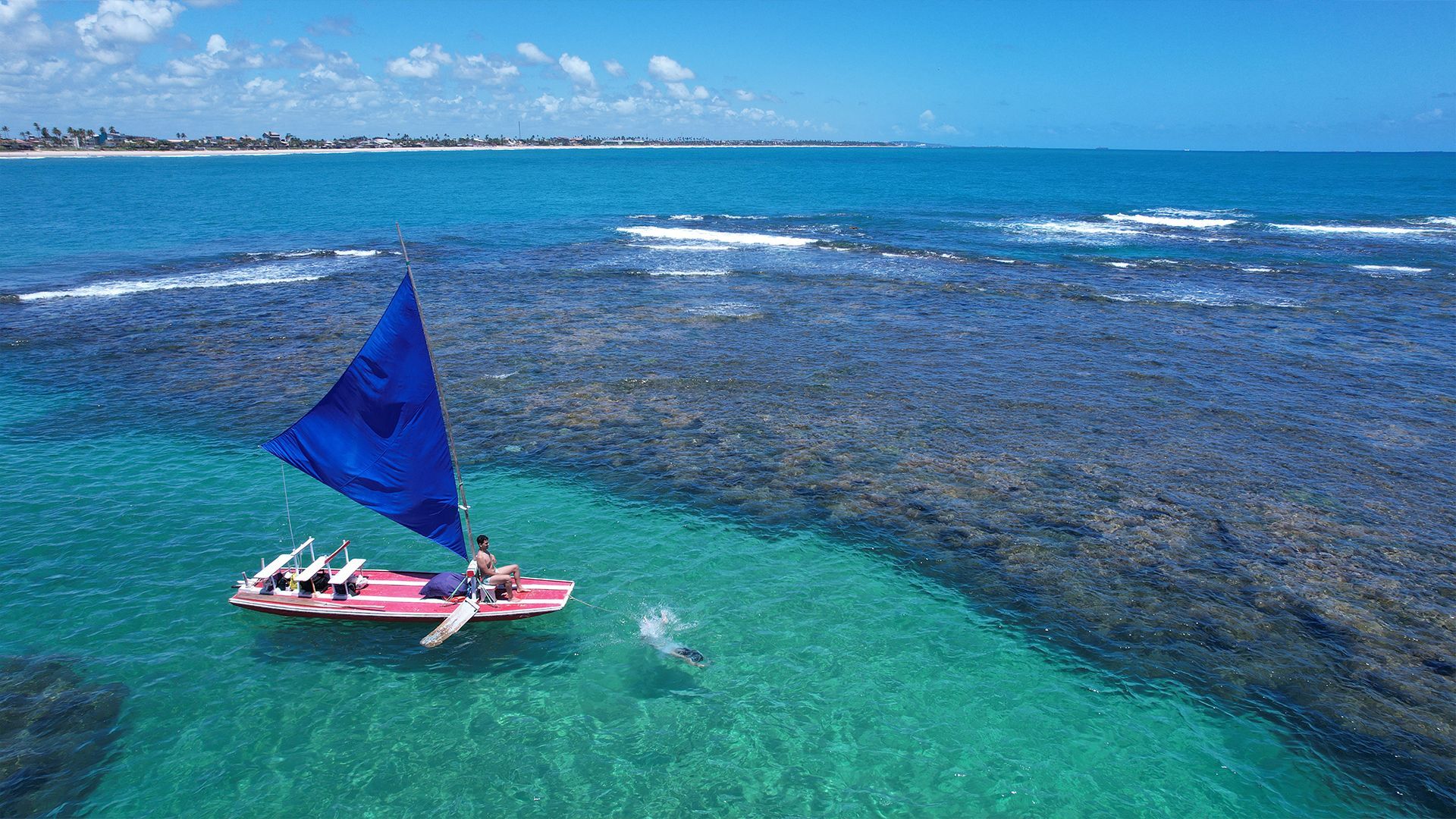 Vista aérea das pisicnas naturais de Porto de Galinhas. Uma jangada de vela azul está em foco.