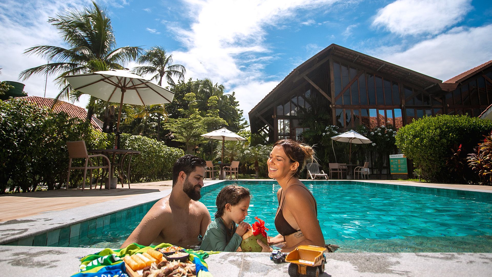 Pai, mãe e filho pequeno curtem a piscina do Armação Resort. Eles estão sorrindo e a criança toma uma água de coco. Na frente deles, na borda da piscina tem uma bandeja de petiscos e ao fundo a estrutura do resort com cadeiras, guarda-sóis e o céu azul e ensolarado. Eles também estão cercados de muito verde, com plantas do jardim.