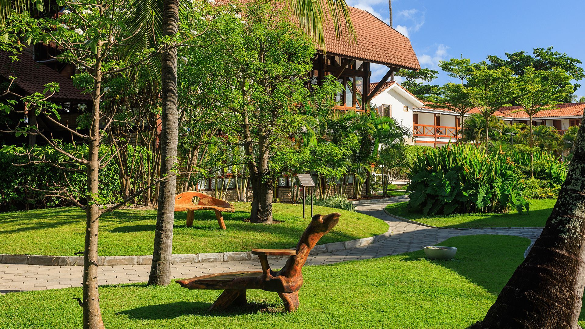 Jardim bem cuidado com gramado verde, árvores tropicais e arbustos. Há caminhos de pedra que percorrem o espaço e bancos de madeira rústicos espalhados pelo local. O céu está azul, sugerindo um dia ensolarado no Armação Resort.