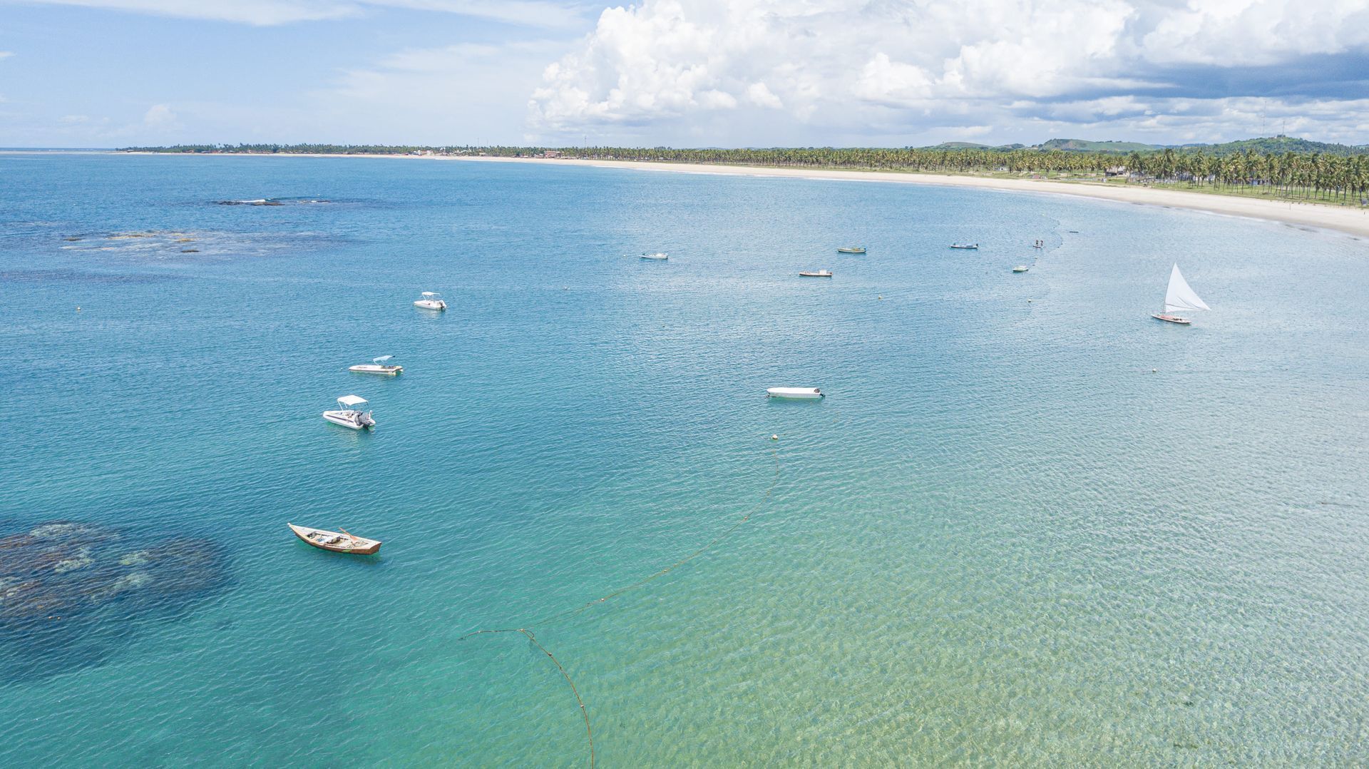 Vista aérea das Piscinas Naturais de Porto de Galinhas, com águas calmas e cristalinas em tons de azul e verde com diversos barcos ancorados próximos à costa.