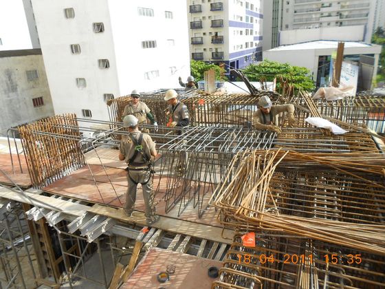 Trabalhadores da construção civil construindo com vergalhões no telhado de um arranha-céu. Alguns usam cintos de segurança. Luz do dia intensa.