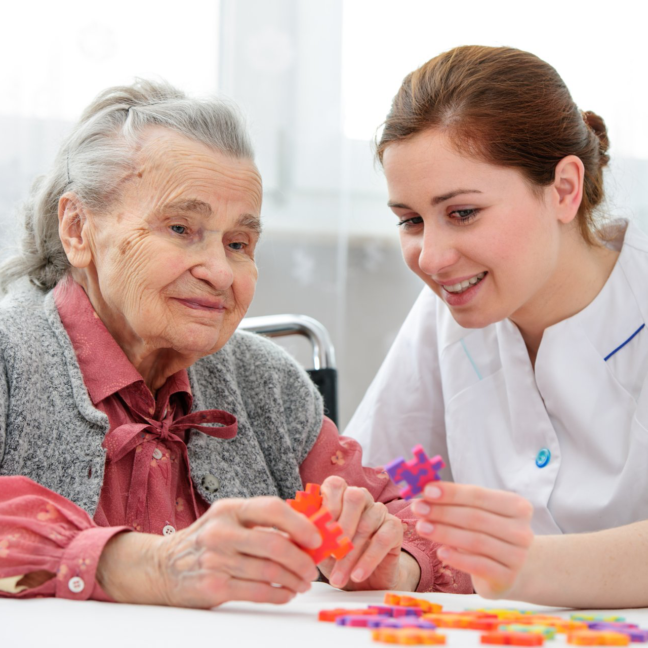 An elderly woman and a caregiver are playing with colorful puzzle pieces at a table. They are both smiling.