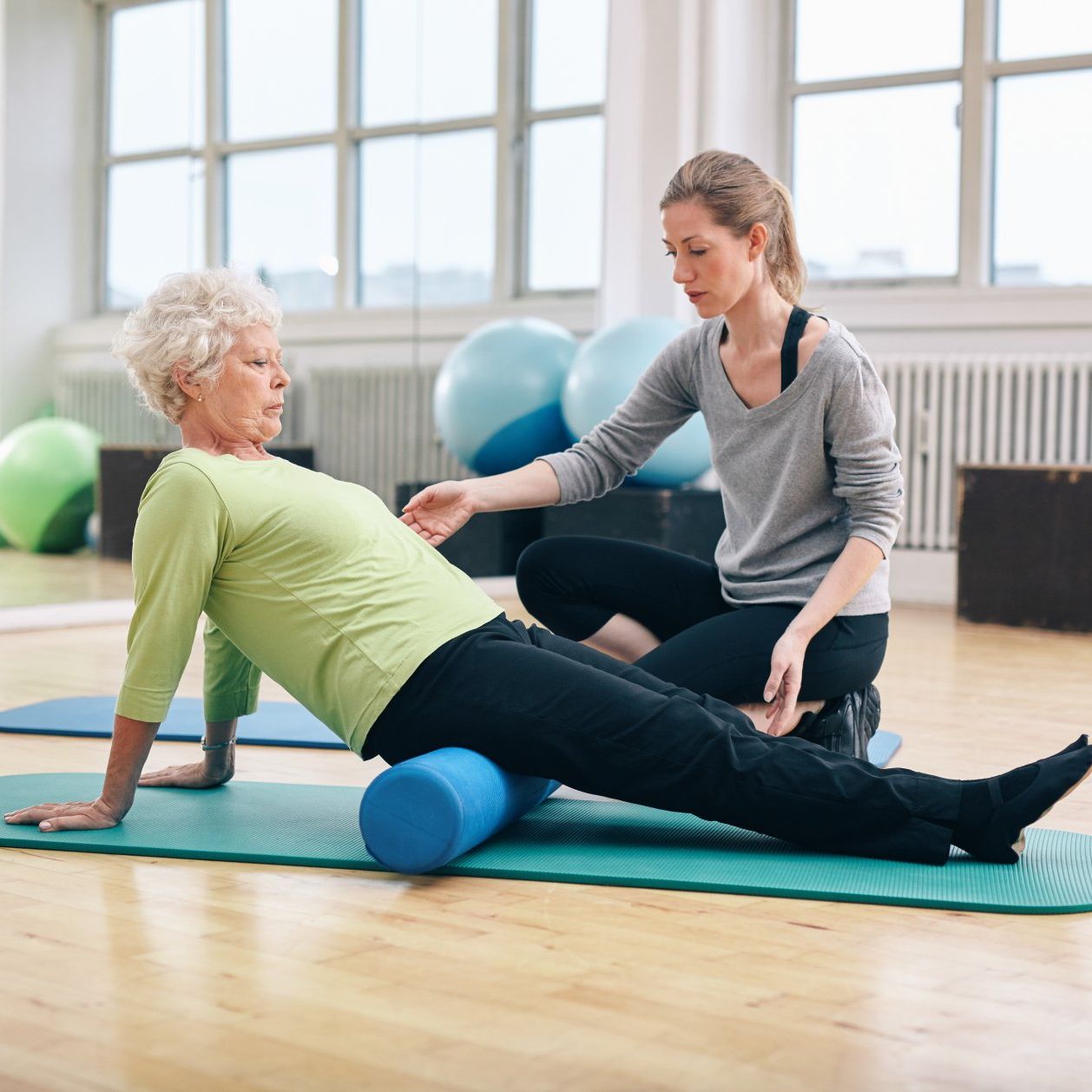 An older woman using a foam roller with a trainer's guidance in a gym. The woman is on a mat with the roller under her thigh.