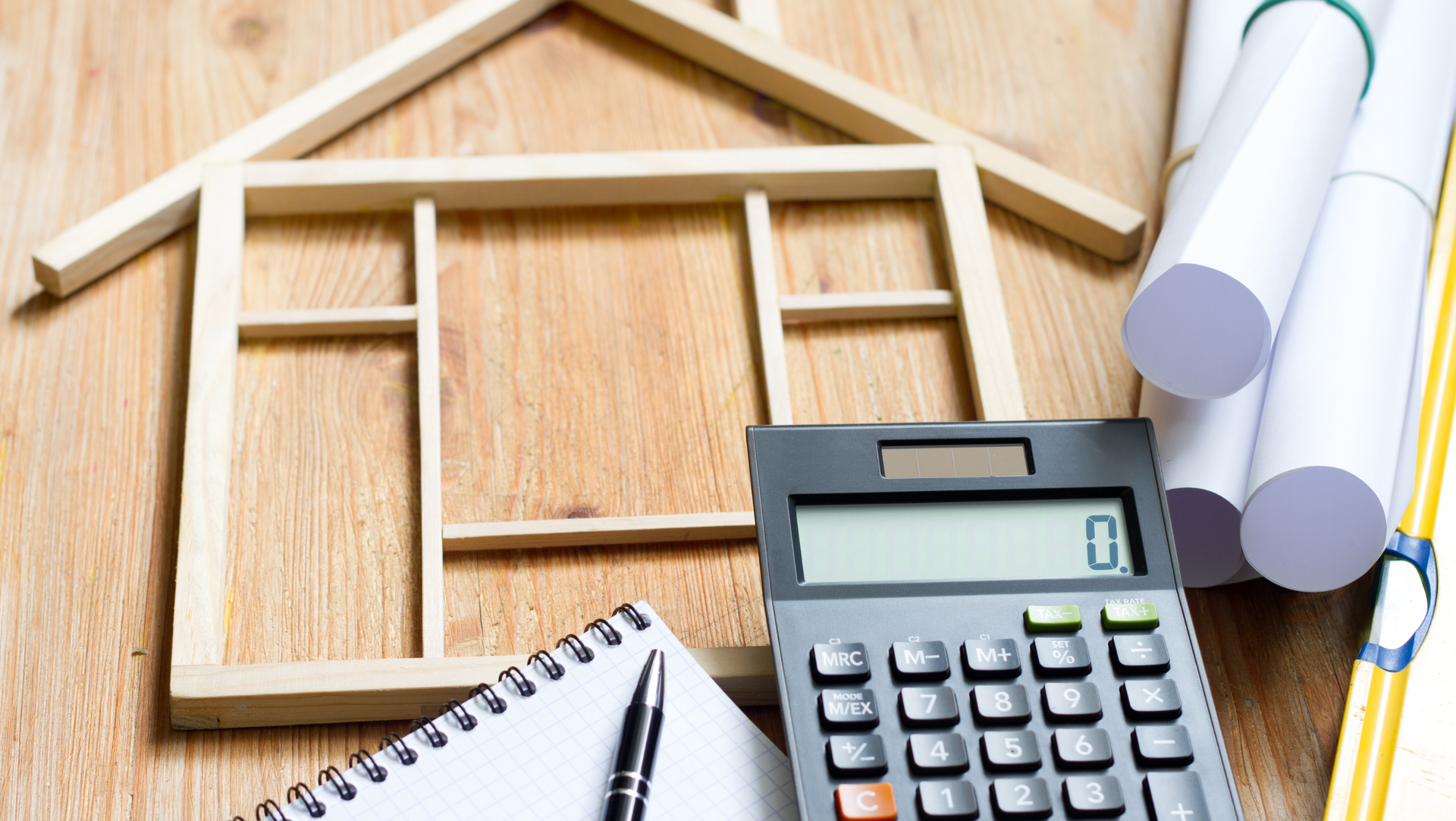 A calculator, notepad, pen, and blueprints sitting on a wooden table next to a model of a house